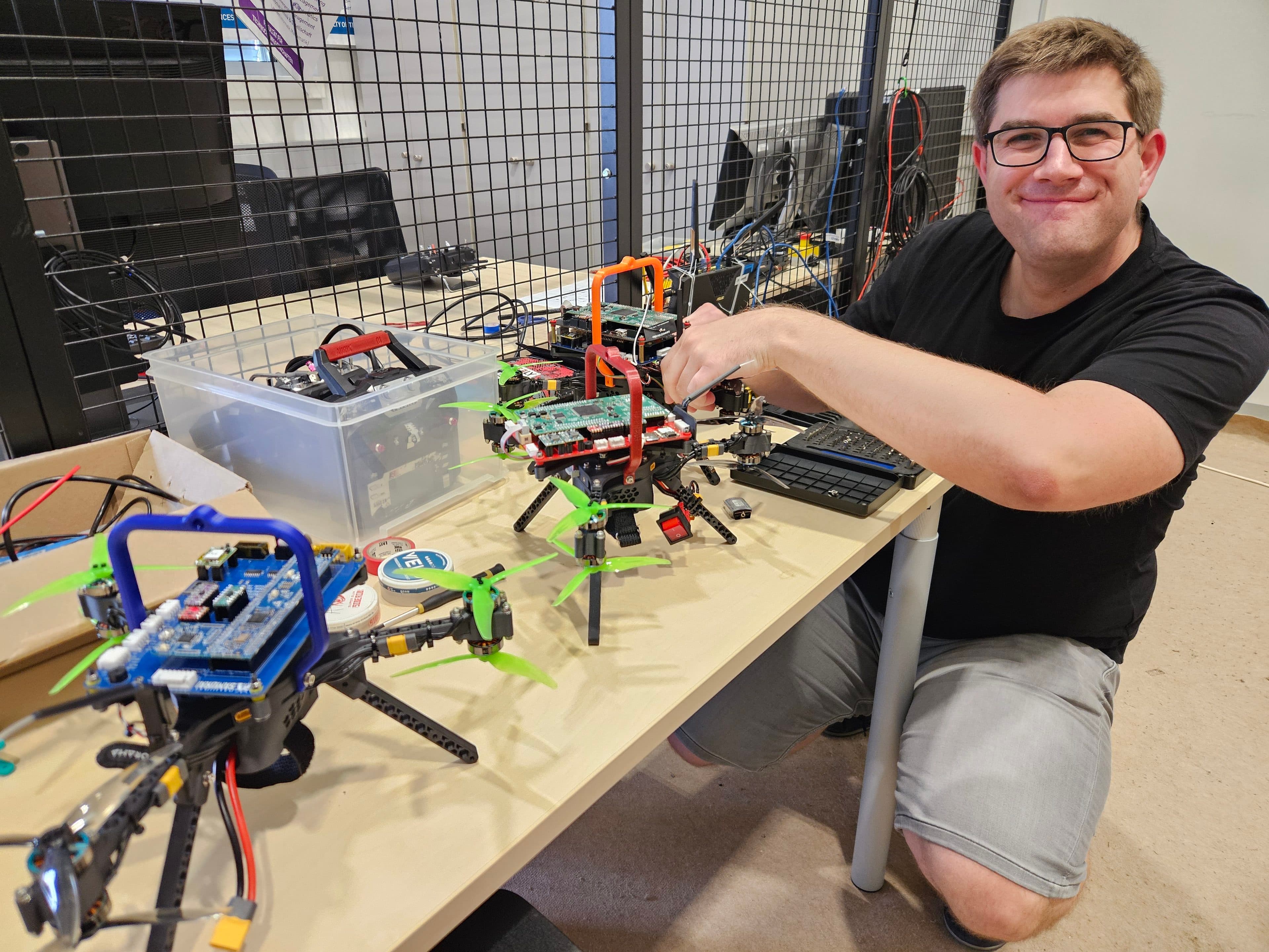 Eine Person mit Brille und schwarzem Hemd lächelt, während sie an einem Schreibtisch in einer Laborumgebung an mehreren kleinen Drohnen mit bunten Propellern arbeitet.