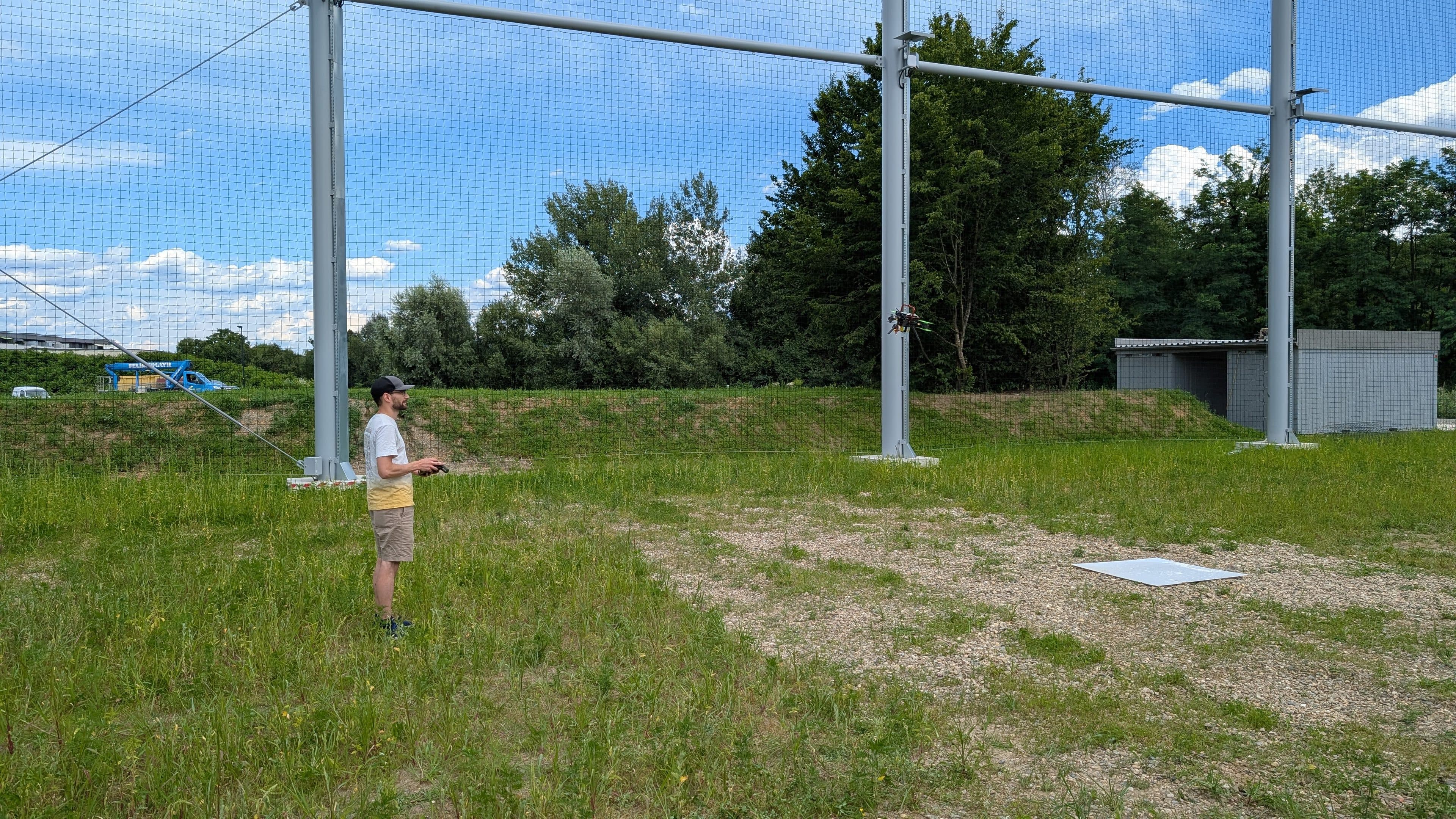 Eine Person steht auf einer grasbewachsenen Freifläche mit Metallkonstruktionen und Netzen und bedient ein ferngesteuertes Gerät. Im Hintergrund sind unter einem teilweise bewölkten Himmel Bäume und ein kleines Gebäude zu sehen.