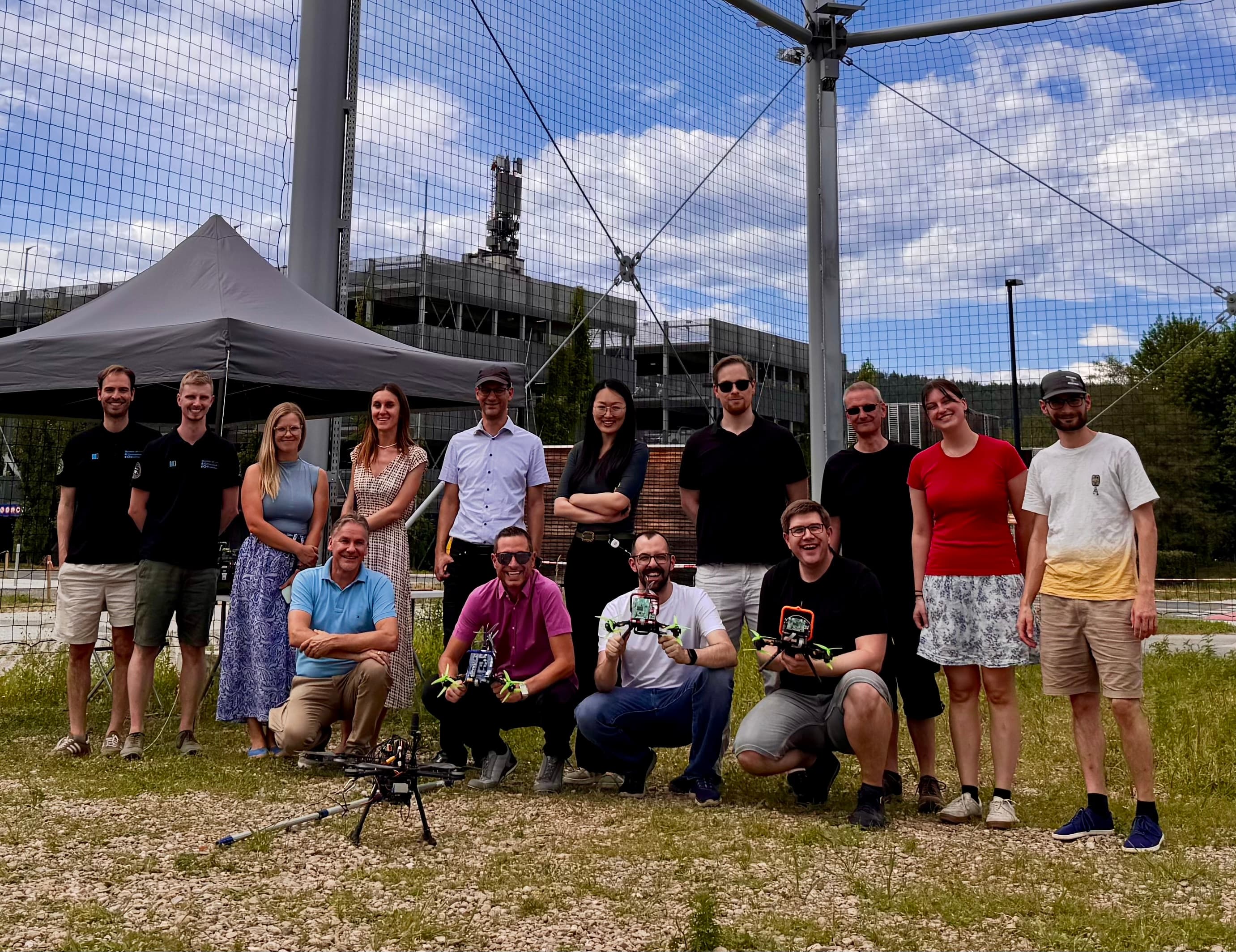 Eine Gruppe von vierzehn Personen, von denen einige stehen und andere knien, posiert an einem sonnigen Tag im Freien in der Nähe einer Drohne unter einem Festzelt, im Hintergrund sind eine eingezäunte Anlage und Bäume zu sehen.