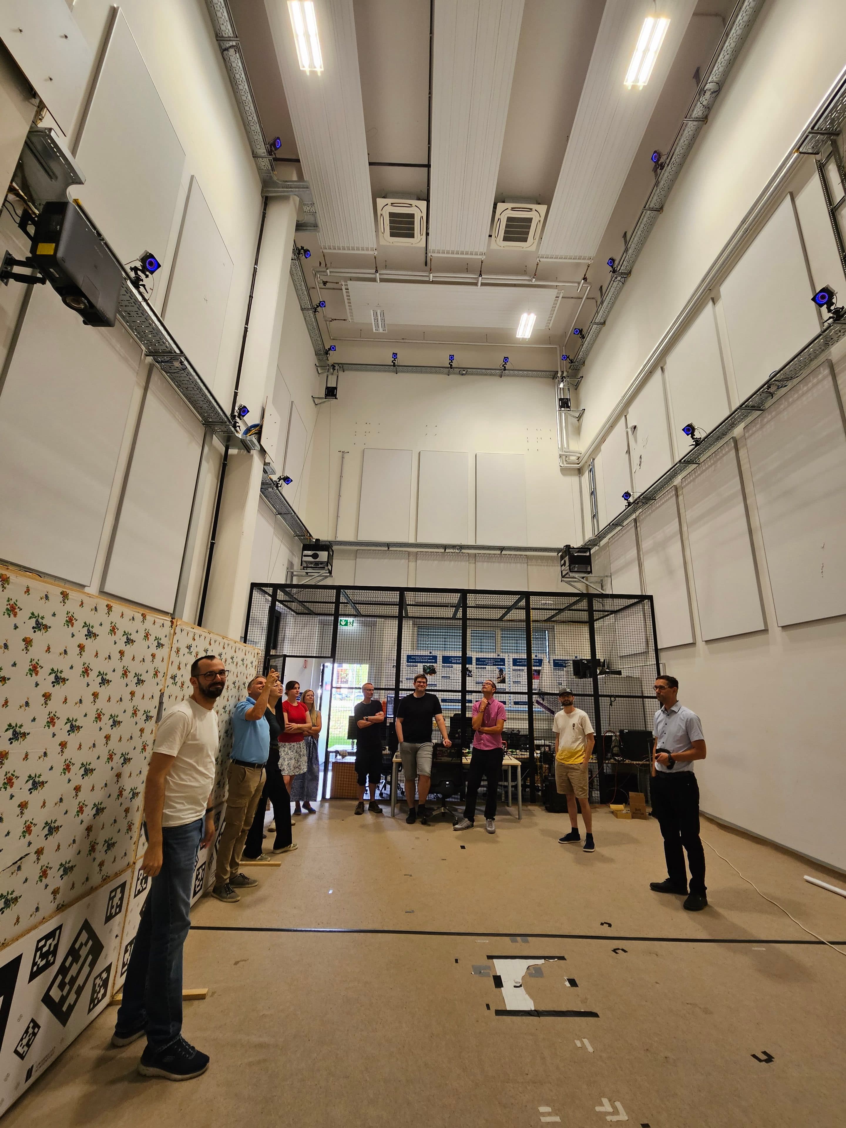 Eine Gruppe von Menschen steht in einem geräumigen Raum mit hoher Decke, an dessen Wänden Motion-Capture-Kameras angebracht sind und im Hintergrund die Ausrüstung aufgestellt ist. Einige unterhalten sich, andere beobachten.