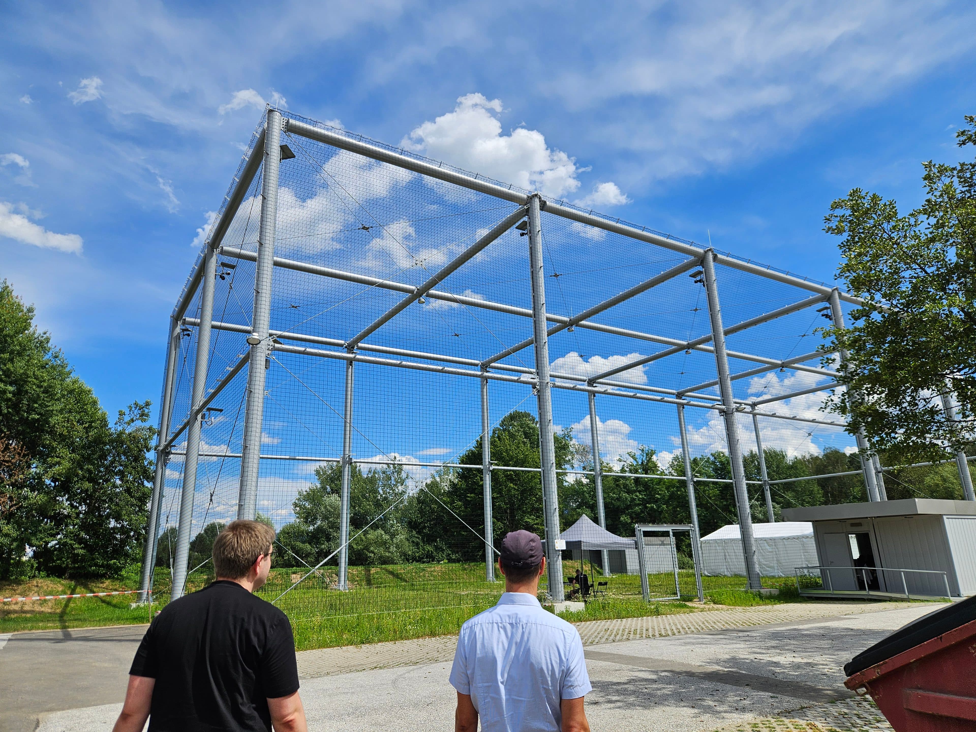 Zwei Menschen stehen vor einer großen Freiluftvoliere aus Metallträgern und Netzgewebe; im Hintergrund sind Bäume und Zelte zu sehen, über ihnen ein blauer Himmel mit vereinzelten Wolken.