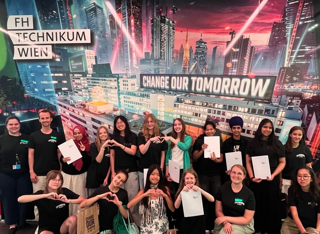 Eine Gruppe von Studierenden posiert in einem Innenraum vor einem Wandbild mit Stadtansicht, auf dem die Schriftzüge „FH Technikum Wien“ und „Change Our Tomorrow“ zu sehen sind. Einige halten Zertifikate in den Händen, viele formen mit ihren Händen ein Herz.