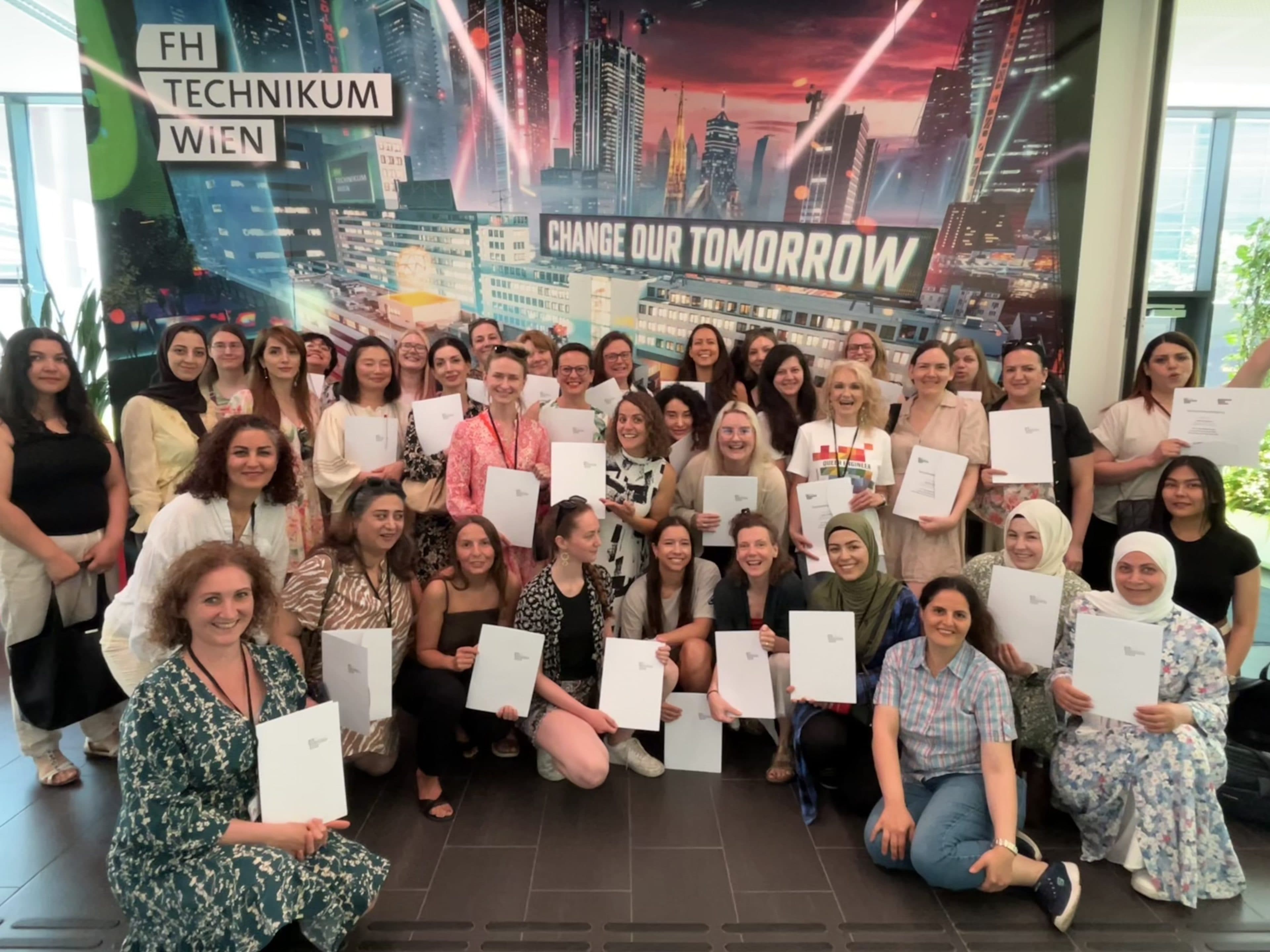 Eine Gruppe von Menschen steht in einem Raum, lächelt und hält Urkunden vor einer farbenfrohen Stadtkulisse in die Kamera, auf der „CHANGE OUR TOMORROW“ und „FH TECHNIKUM WIEN“ zu lesen ist.