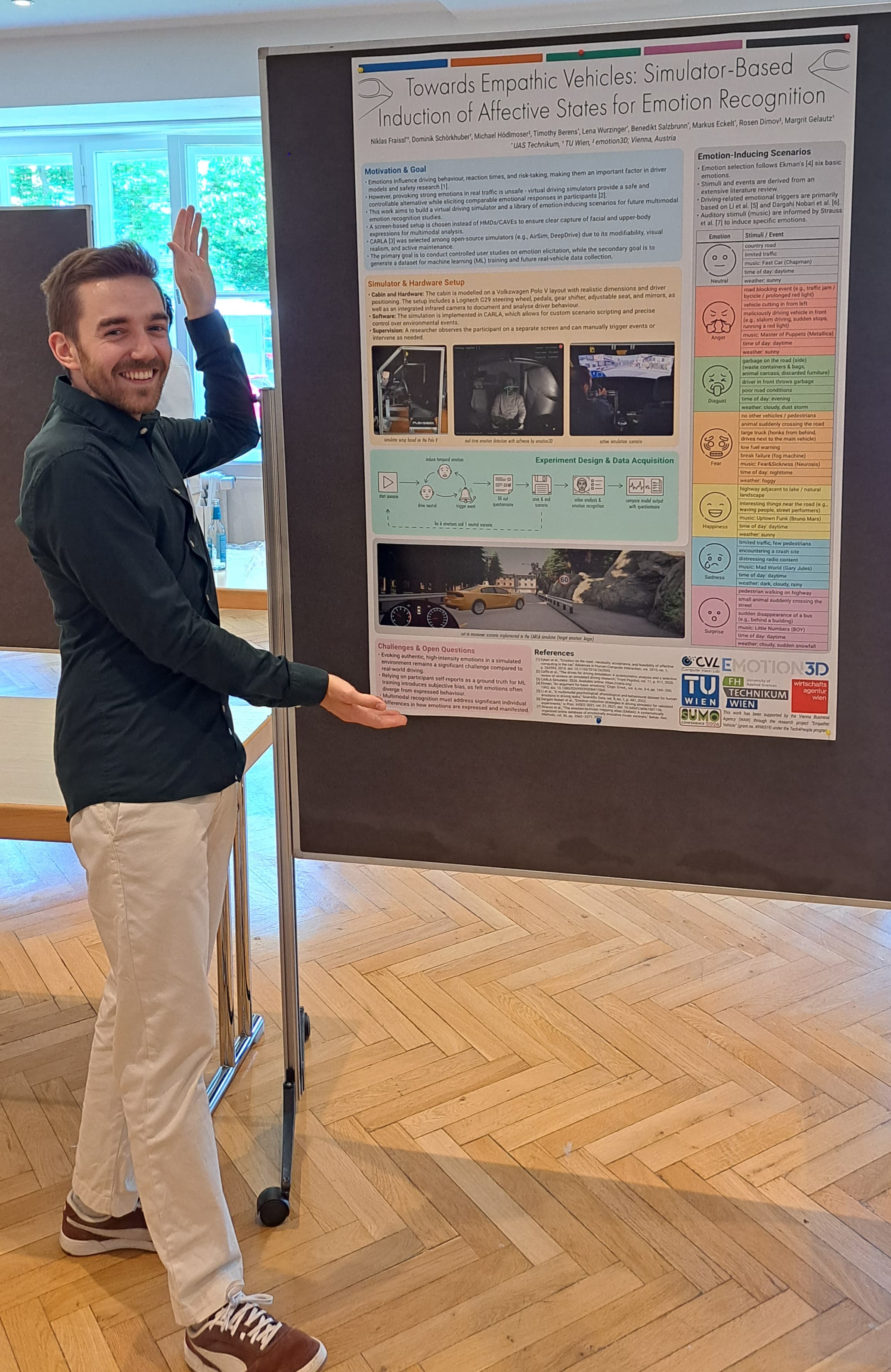 Eine Person steht in einem Raum, lächelt und deutet mit beiden Händen auf ein wissenschaftliches Poster mit dem Titel „Towards Empathic Vehicles“, das an einer Tafel hängt. Der Raum hat Holzböden und große Fenster, durch die man Bäume sehen kann.