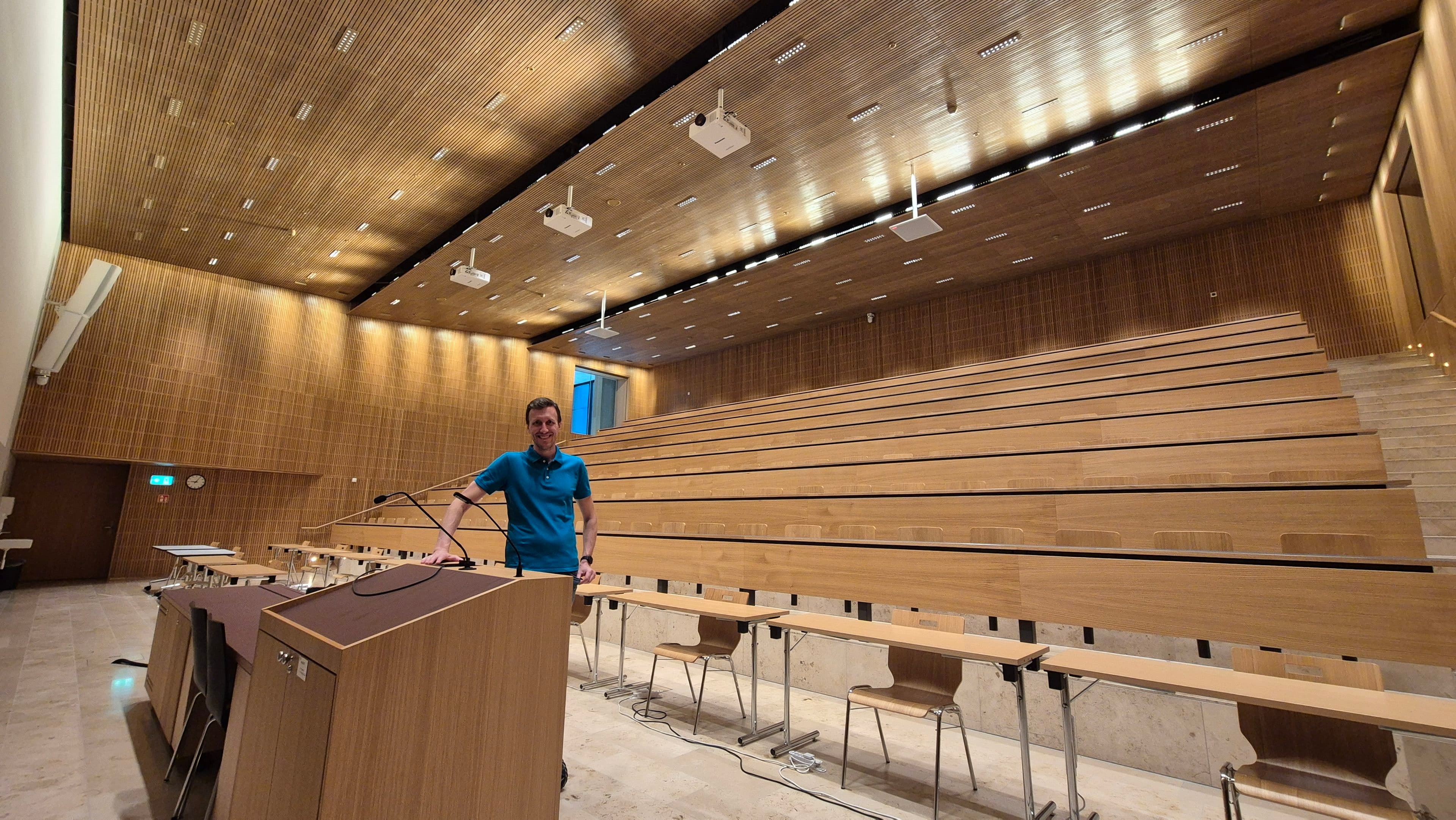 Eine Person steht an einem Rednerpult in einem großen, leeren Hörsaal mit Holzwänden, gestuften Sitzreihen und Deckenleuchten.