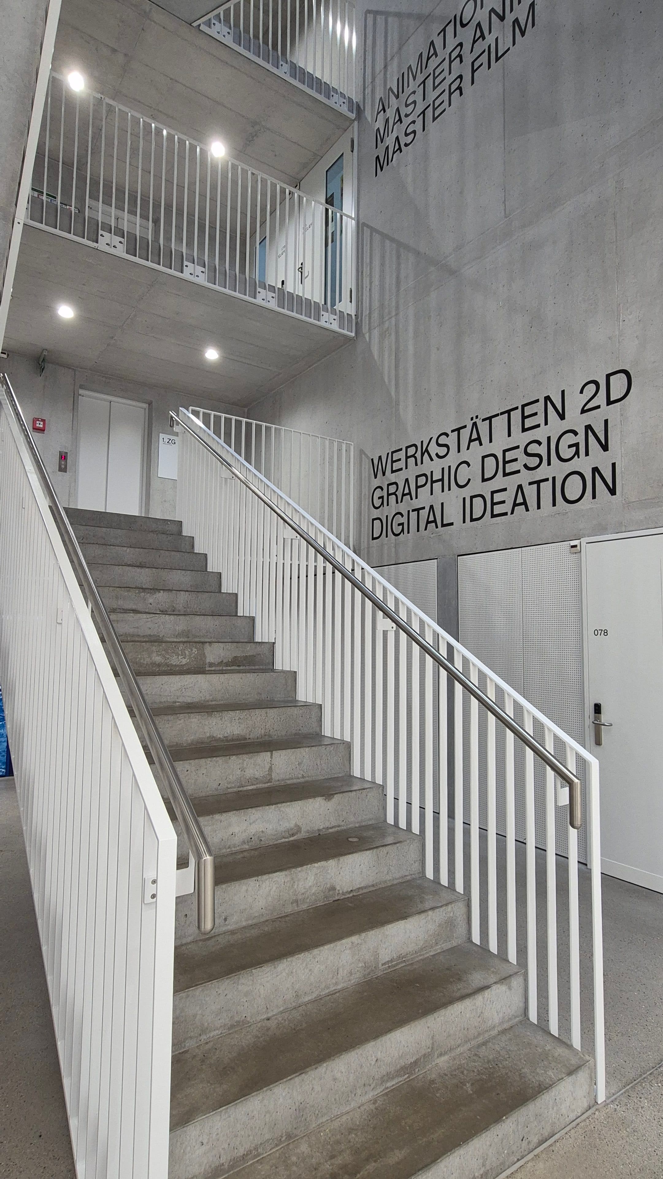 Modernes Treppenhaus mit weißen Geländern und grauen Betonwänden, an denen schwarze Schriftzüge den Weg zu den Räumen für Animation, Masterfilm, 2D-Workshops, Grafikdesign und digitale Ideenfindung weisen.