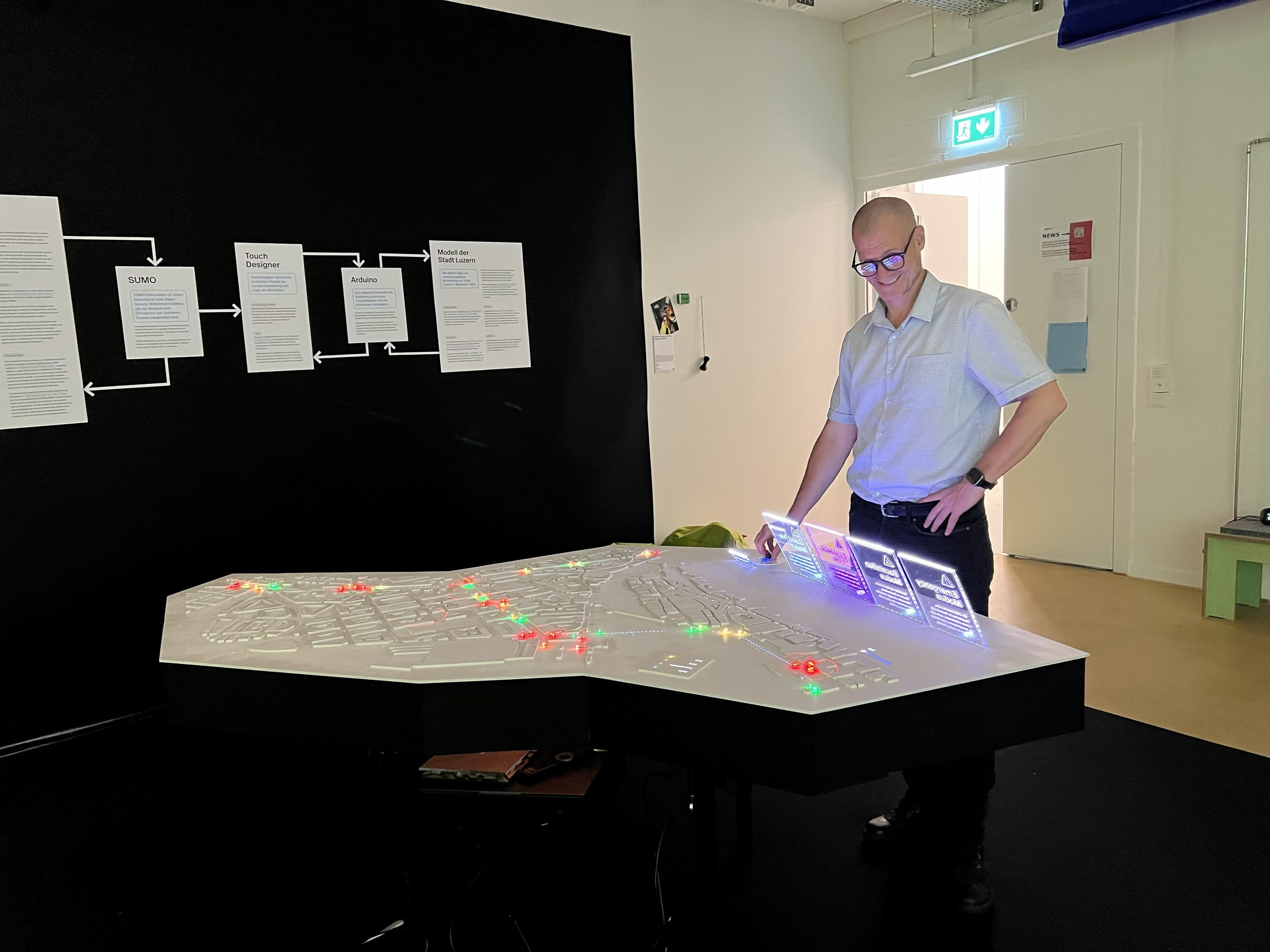 Eine Person steht vor einer beleuchteten Tischkarte mit farbigen Lichtern und interagiert mit einem transparenten Display. Hinter ihr sind Informationsplakate an einer schwarzen Wand angebracht. Der Raum hat weiße Wände und ein beleuchtetes Ausgangsschild.