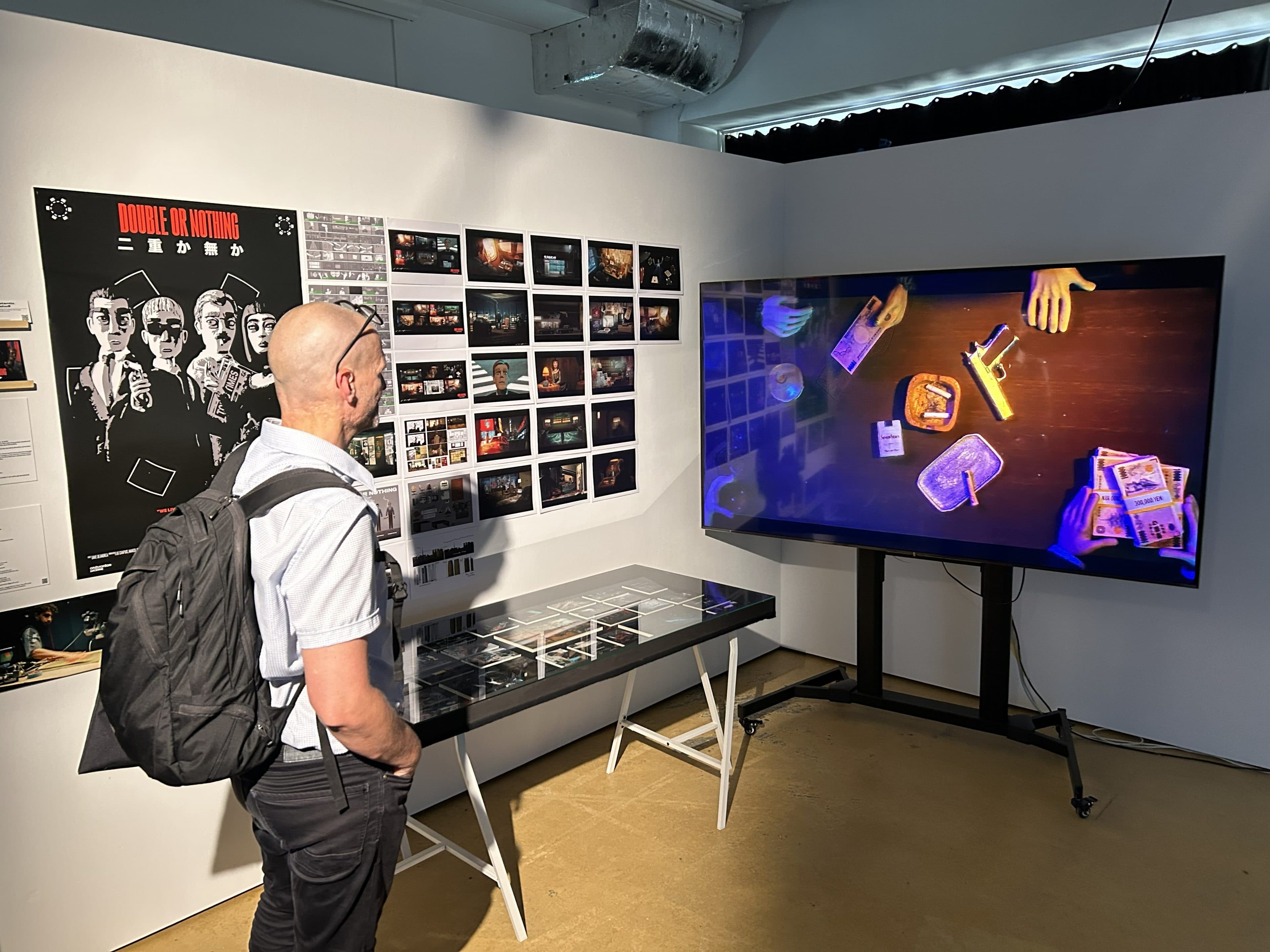 Ein Mann mit einem Rucksack betrachtet in einem modernen Ausstellungsraum eine digitale Anzeige, auf der Hände, Karten und Geld zu sehen sind, neben einer Wand mit Plakaten und Storyboard-Bildern.
