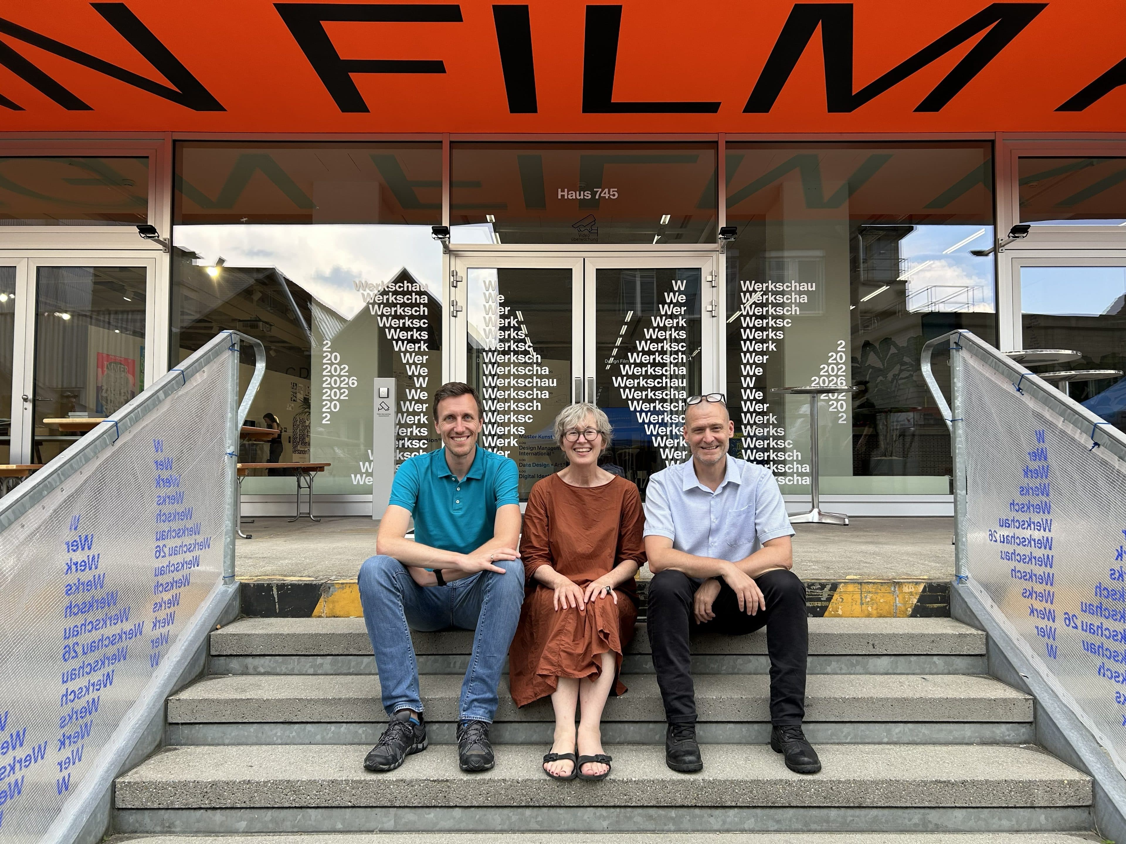 Drei Personen sitzen nebeneinander auf einer Freitreppe und lächeln in die Kamera. Hinter ihnen befindet sich ein gläserner Eingangsbereich mit Schriftzügen an den Türen und einem orangefarbenen Vordach, auf dem „FILM“ steht.