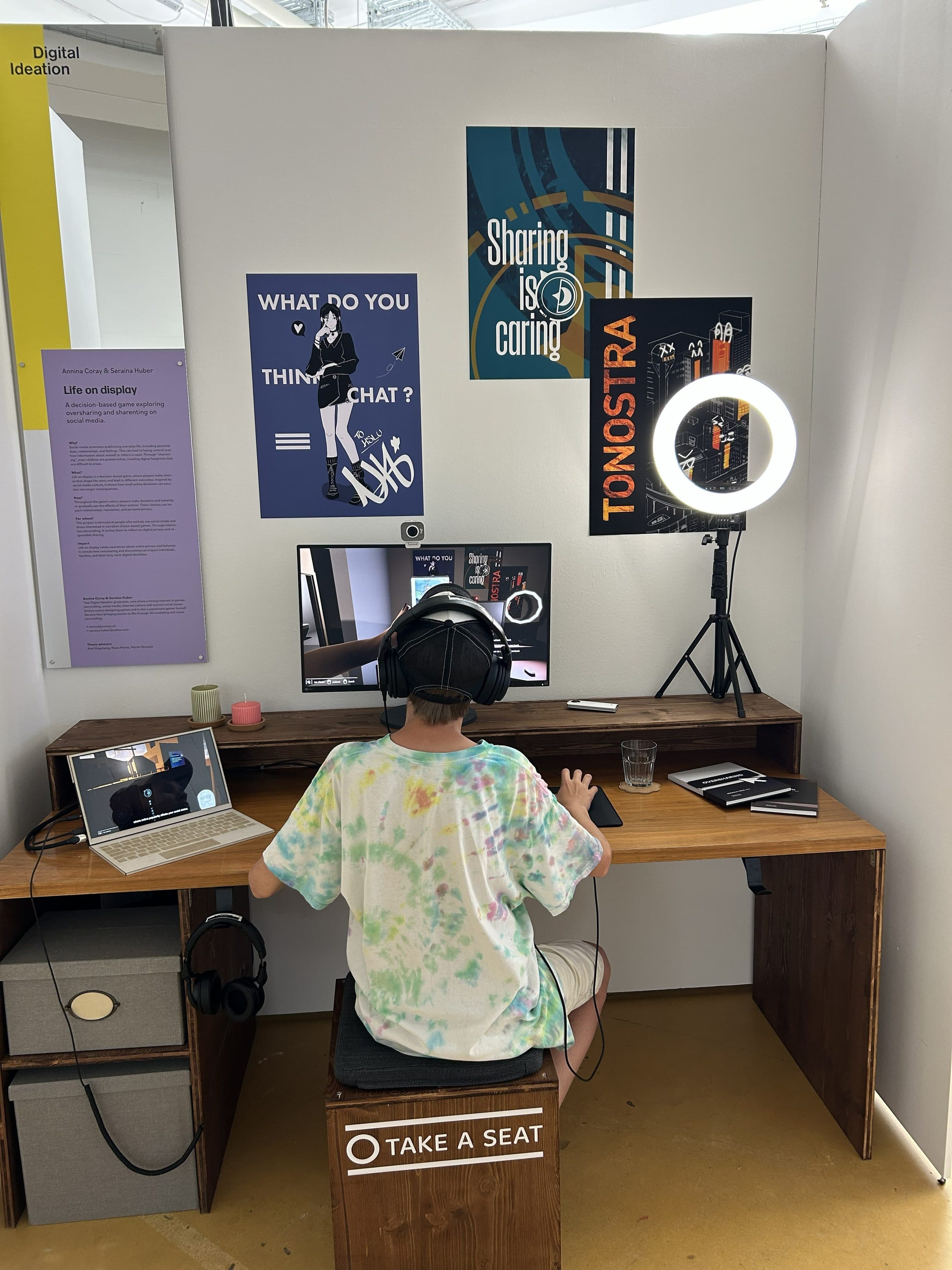 Eine Person mit Kopfhörern sitzt an einem Schreibtisch, auf dem ein Computer, ein Laptop und ein Ringlicht stehen. An der Wand hängen Plakate und Zettel, darunter „WHAT DO YOU THINK—CHAT?“ und „Sharing is caring“. Auf einem Schild steht: „TAKE A SEAT…“.