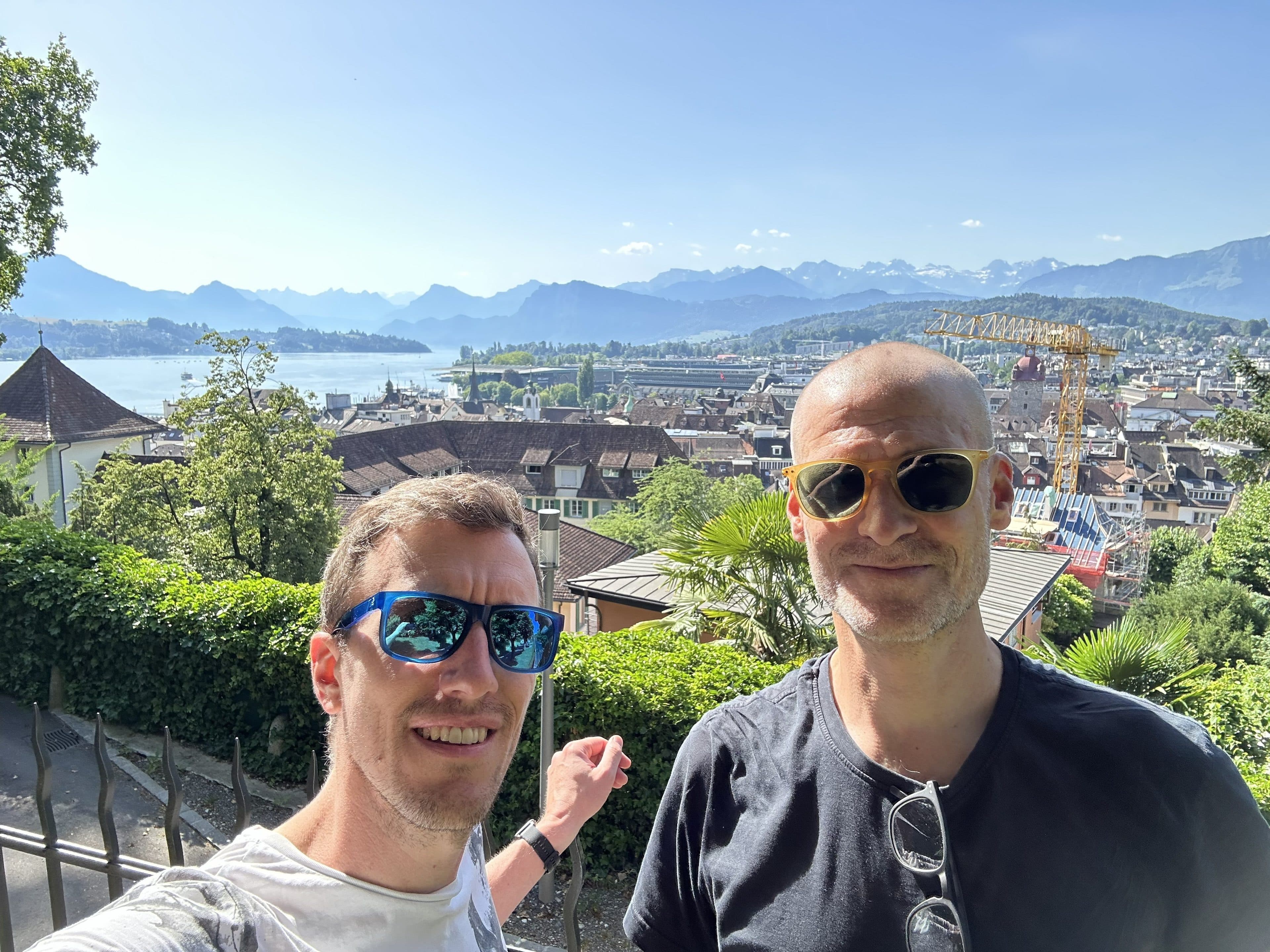 Zwei Männer mit Sonnenbrillen lächeln in die Kamera, während im Hintergrund unter einem strahlend blauen Himmel eine malerische Aussicht auf eine Stadt, einen See und ferne Berge zu sehen ist.