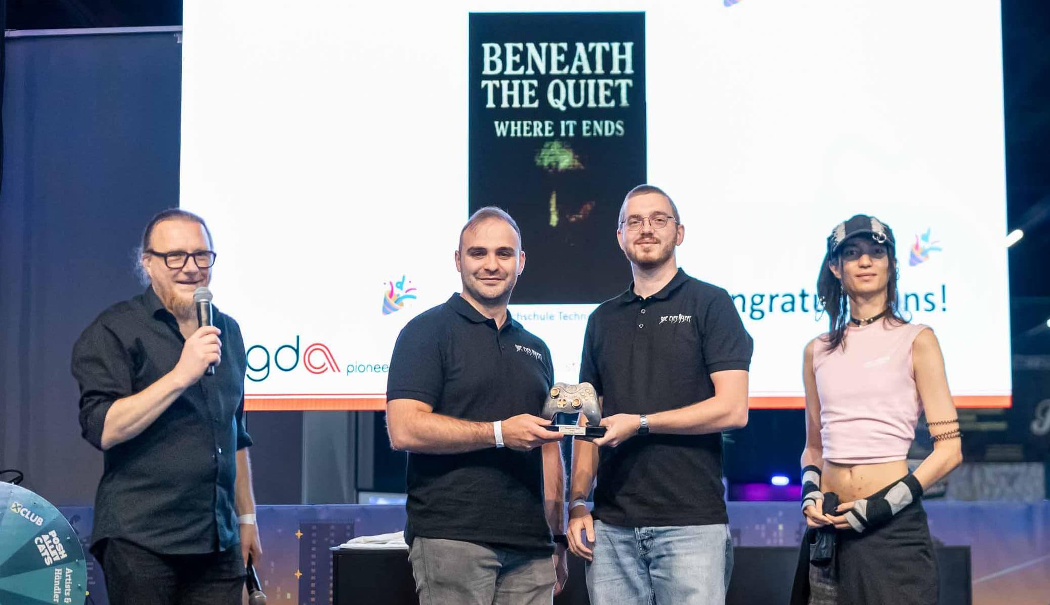 Bei einer Preisverleihung stehen vier Personen auf der Bühne. Zwei Männer in der Mitte halten eine Trophäe in den Händen. Auf einer Leinwand hinter ihnen sind die Schriftzüge „Beneath the Quiet Where It Ends“ und „Gewinner des Talent Award 2023“ zu sehen.