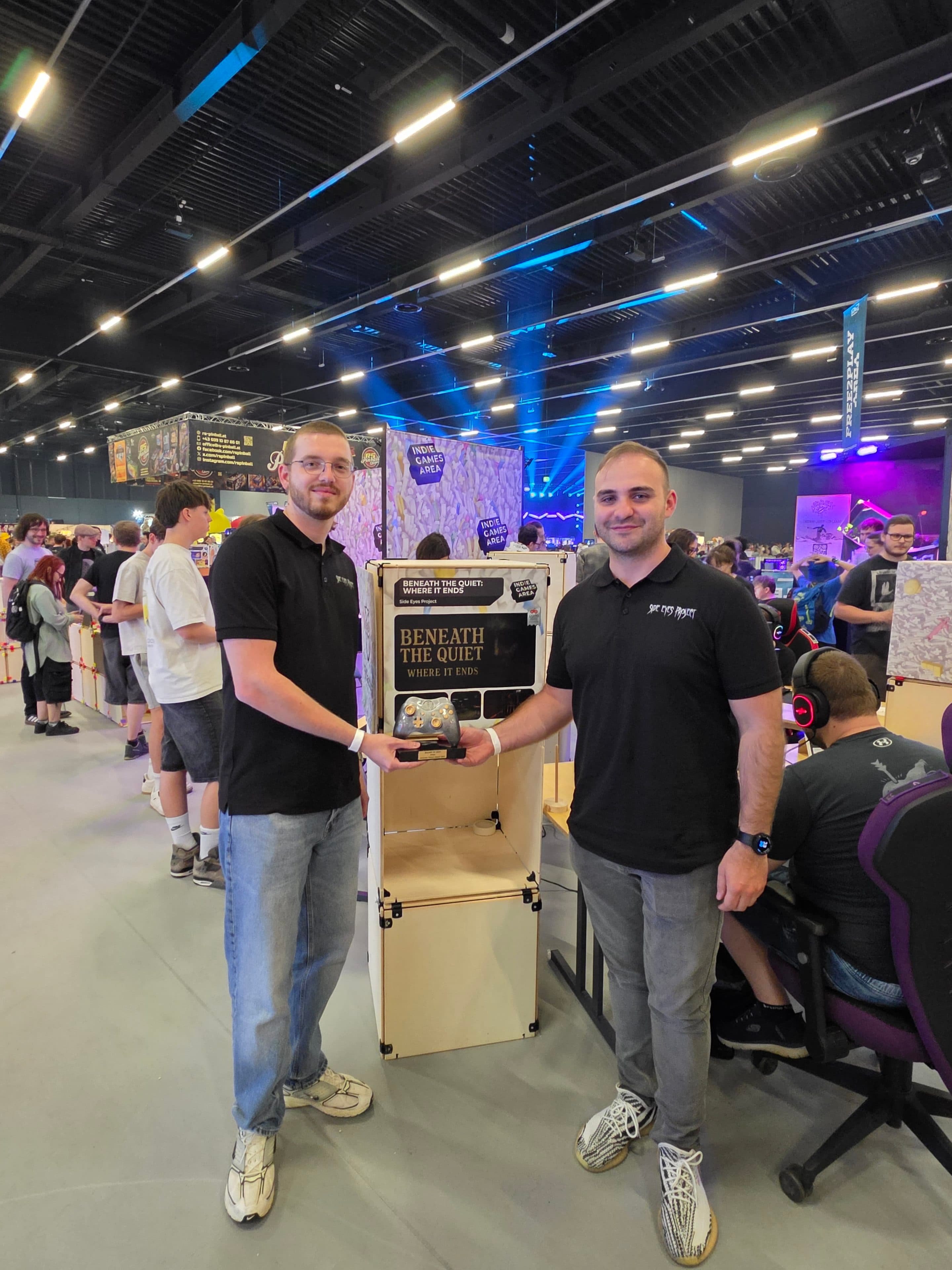 Zwei Männer stehen in einem Raum und halten eine Trophäe für „Beneath The Quiet“, den Gewinner des Jahres 2023 bei einer Gaming-Veranstaltung, in den Händen. Im Hintergrund haben sich Menschen unter grellem Licht um Computer versammelt.