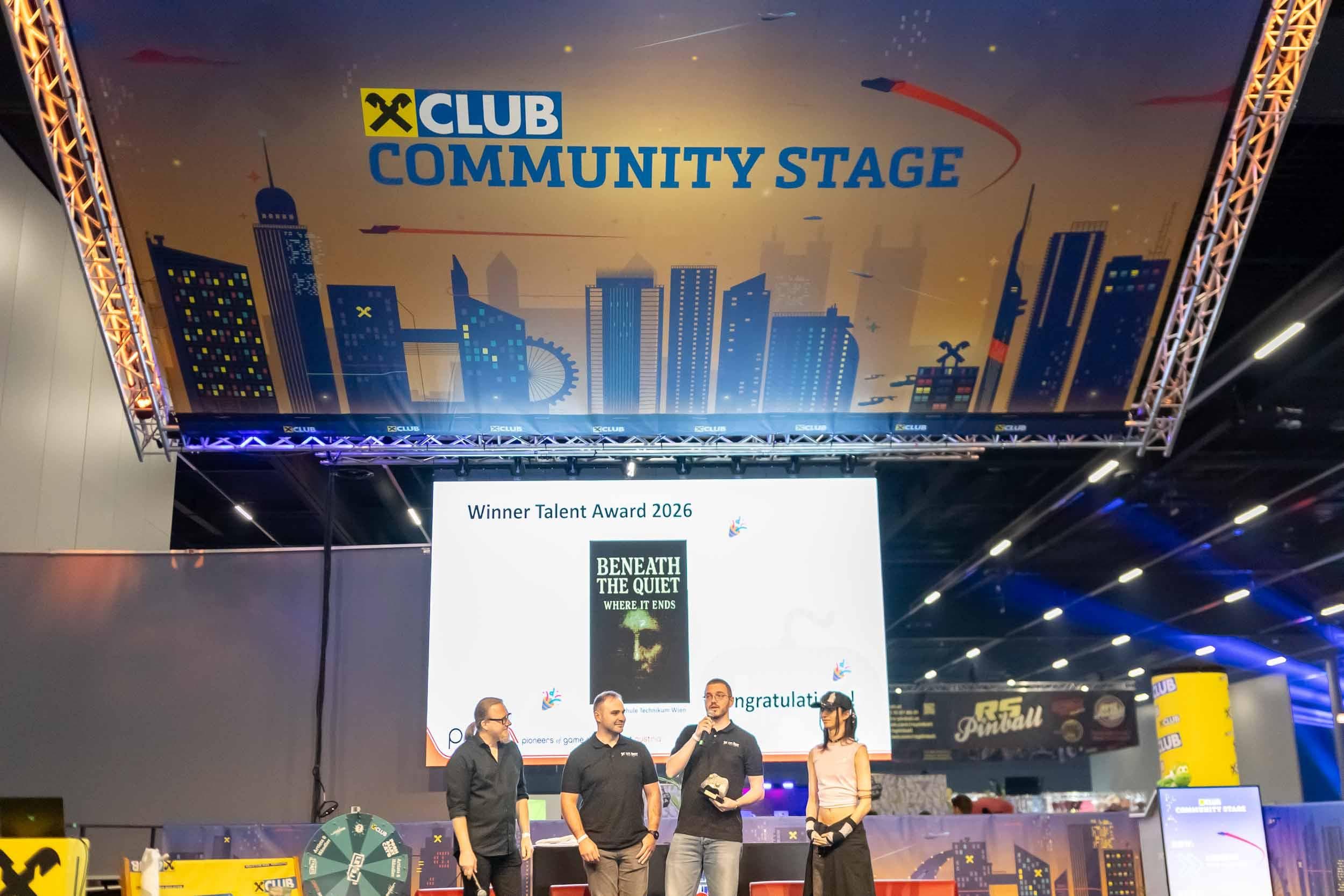 Vier Personen stehen auf einer Bühne bei einer Preisverleihung für Talente, über ihnen hängt ein großes „Community Stage“-Banner, und im Hintergrund ist auf einer Leinwand der Titel „Beneath the Quiet“ zu sehen. Man erkennt grelle Scheinwerfer und die Kulisse einer Veranstaltung in einem Saal.