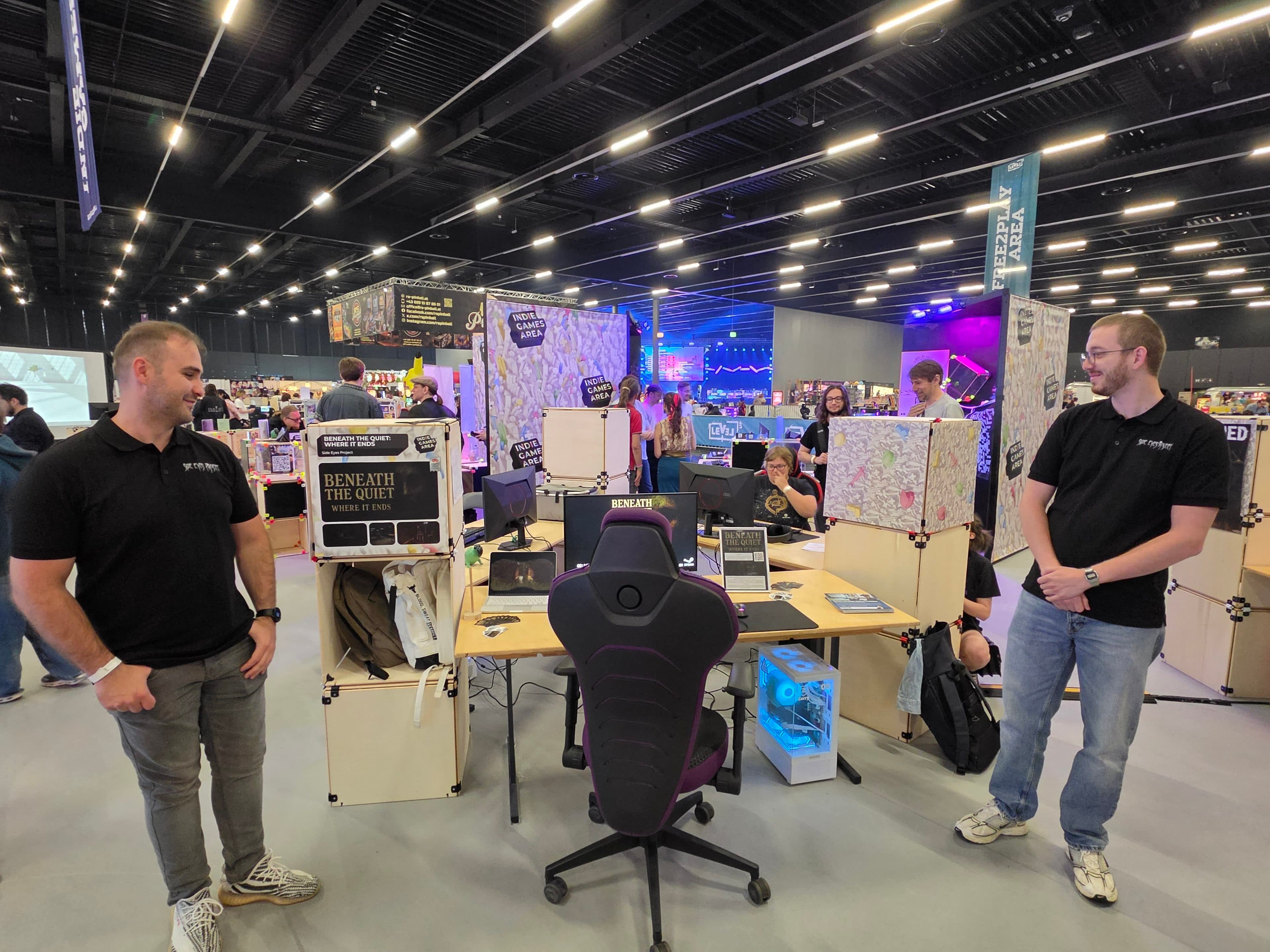 Zwei Männer in schwarzen Hemden stehen in einem Kongresszentrum neben einem Stand mit Karten, Schildern und Bildschirmen. Hinter dem Stand, auf dem „Beneath the Quiet“ zu sehen ist, sitzen oder stehen mehrere Personen. Helle Lichter erhellen den großen Veranstaltungsraum.