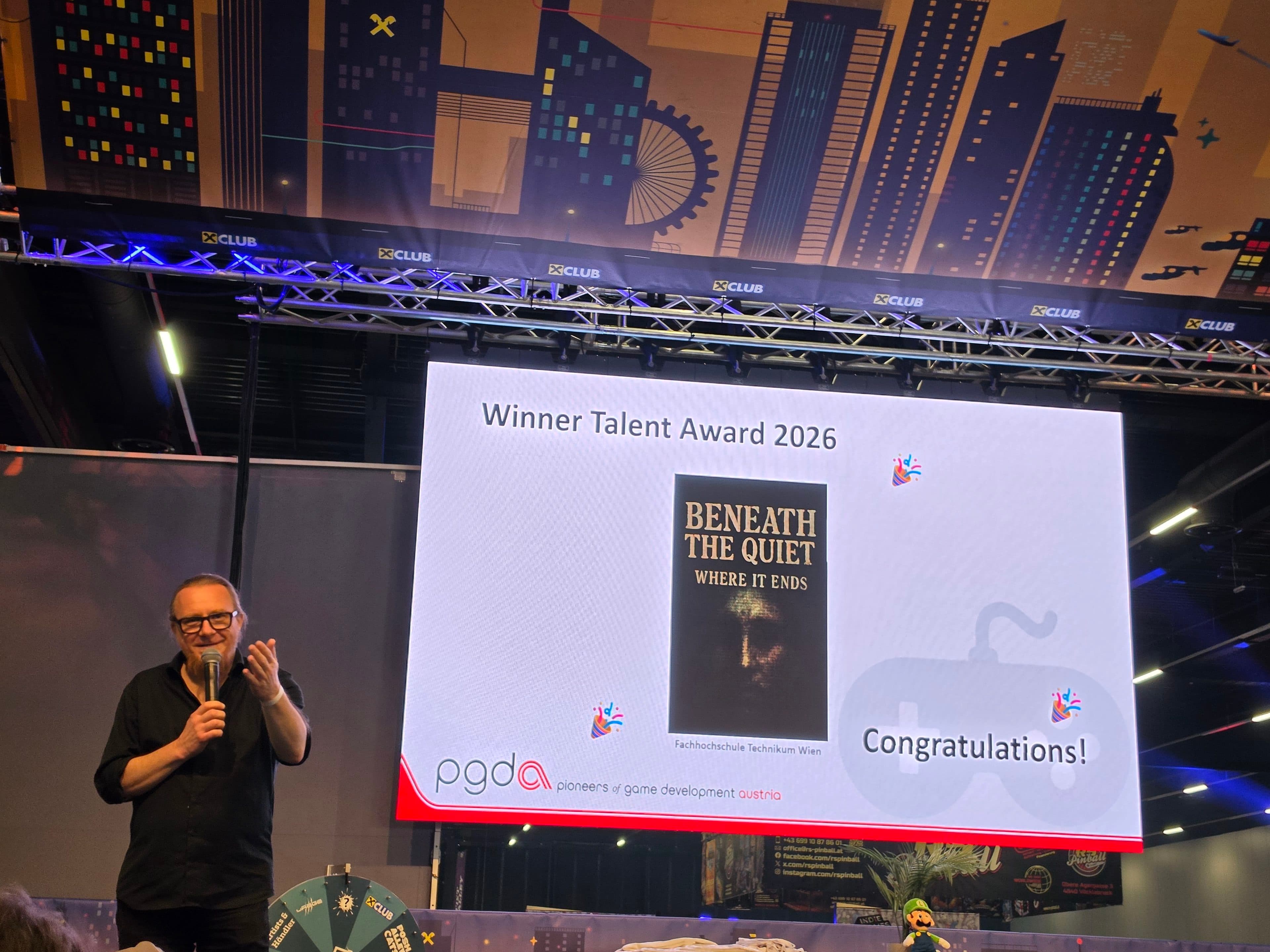 Ein Mann spricht auf der Bühne mit einem Mikrofon. Hinter ihm ist auf einer Leinwand die Auszeichnung „Winner Talent Award 2026“ für „Beneath the Quiet: Where It Ends“ zu sehen. Das PGDA-Logo und die Aufschrift „Herzlichen Glückwunsch!“ erscheinen auf der Leinwand.