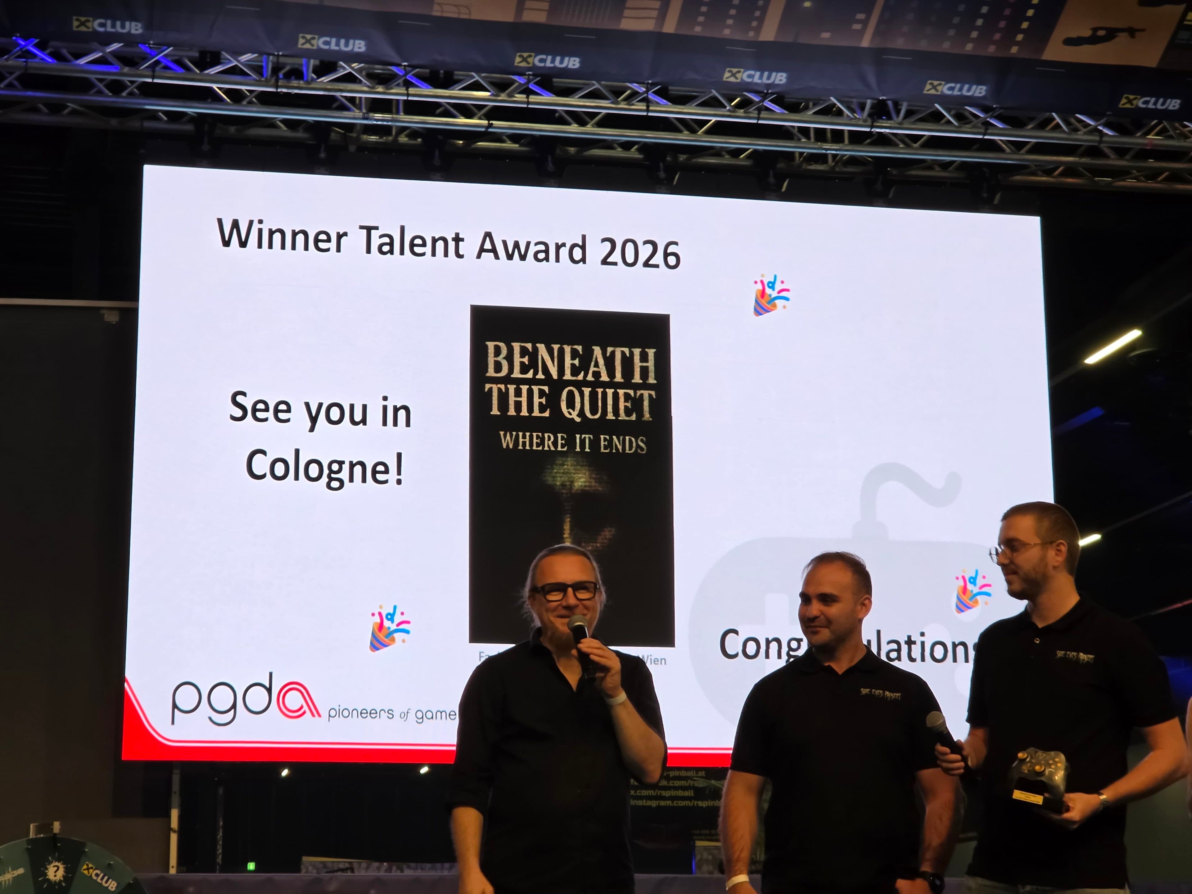 Drei Personen stehen mit Mikrofonen auf der Bühne. Hinter ihnen ist auf einer großen Leinwand der Schriftzug „Winner Talent Award 2026“ zu sehen, zusammen mit einem Buchcover mit dem Titel „Beneath The Quiet: Where It Ends“ und der Botschaft „Wir sehen uns in Köln!“.
