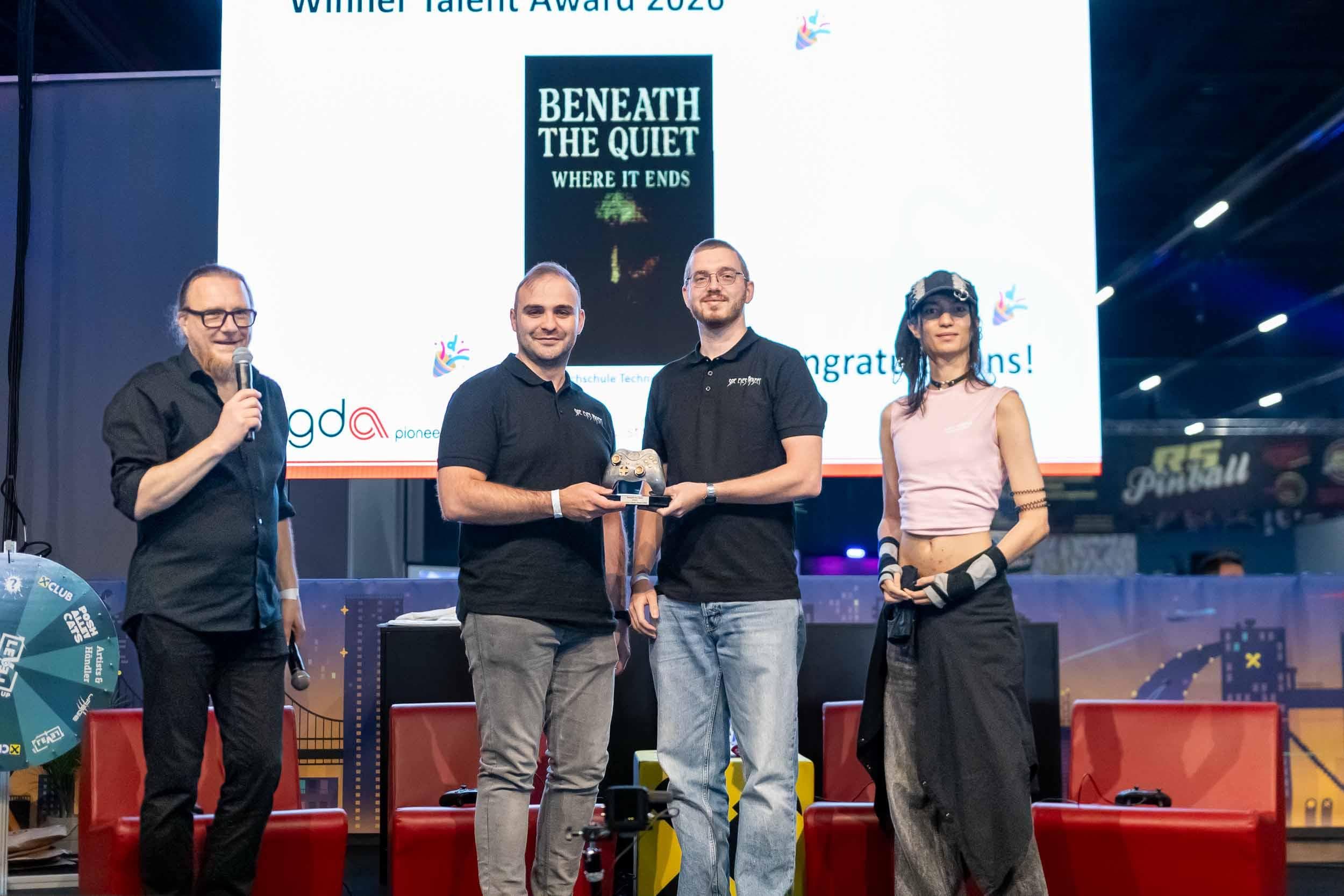 Bei einer Preisverleihung stehen vier Personen auf der Bühne. Zwei Personen in der Mitte halten eine Trophäe in den Händen. Auf einer Leinwand hinter ihnen ist zu lesen: „Gewinner des Talent Award 2023 – Beneath The Quiet: Where It Ends…“.