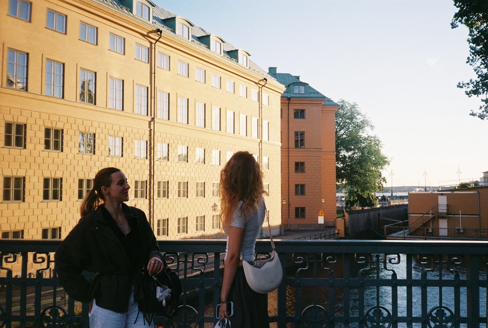 Zwei Frauen stehen auf einer Brücke über einem Kanal und unterhalten sich, im Hintergrund sind große gelbe Gebäude und Bäume zu sehen, über ihnen ein teilweise wolkenloser Himmel.