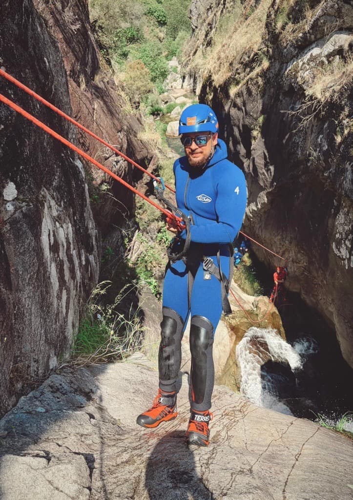 Eine Person in einem blauen Neoprenanzug, mit Helm und roten Stiefeln seilt sich an roten Kletterseilen einen felsigen Canyon hinab, unter dem ein Bach fließt. Im Hintergrund ist ein weiterer Kletterer zu sehen.
