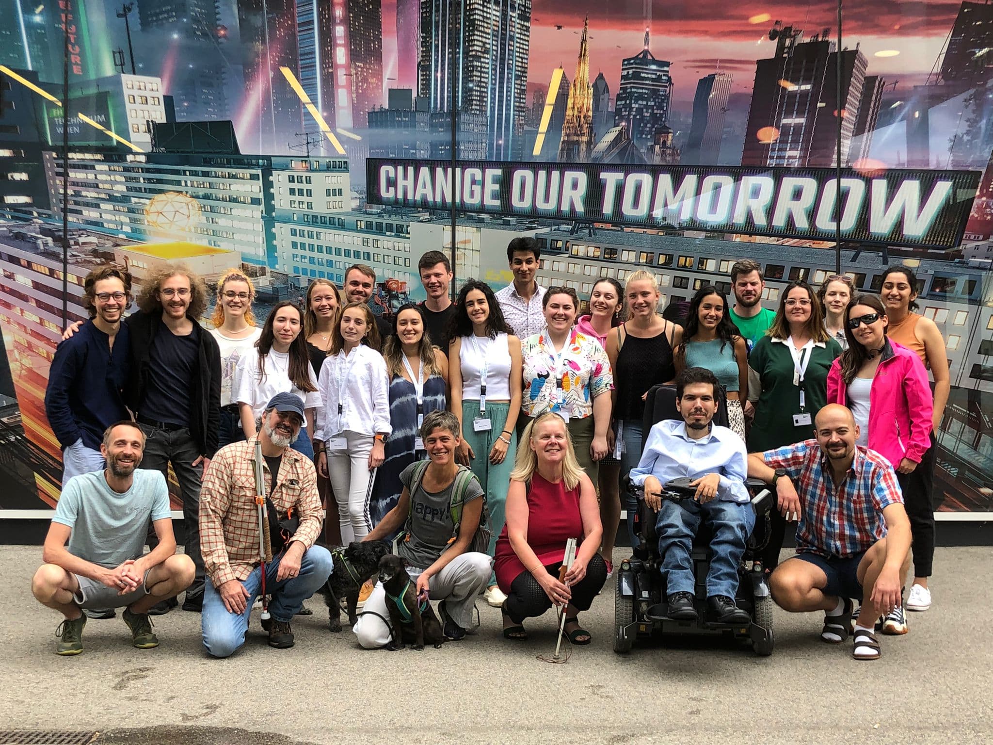 Eine Gruppe von 26 Menschen unterschiedlichster Herkunft posiert lächelnd vor einem Wandgemälde mit Stadtmotiv, auf dem im Hintergrund die Worte „CHANGE OUR TOMORROW“ zu sehen sind. Eine Person sitzt im Rollstuhl, und die Gruppe wirkt fröhlich und freundlich.