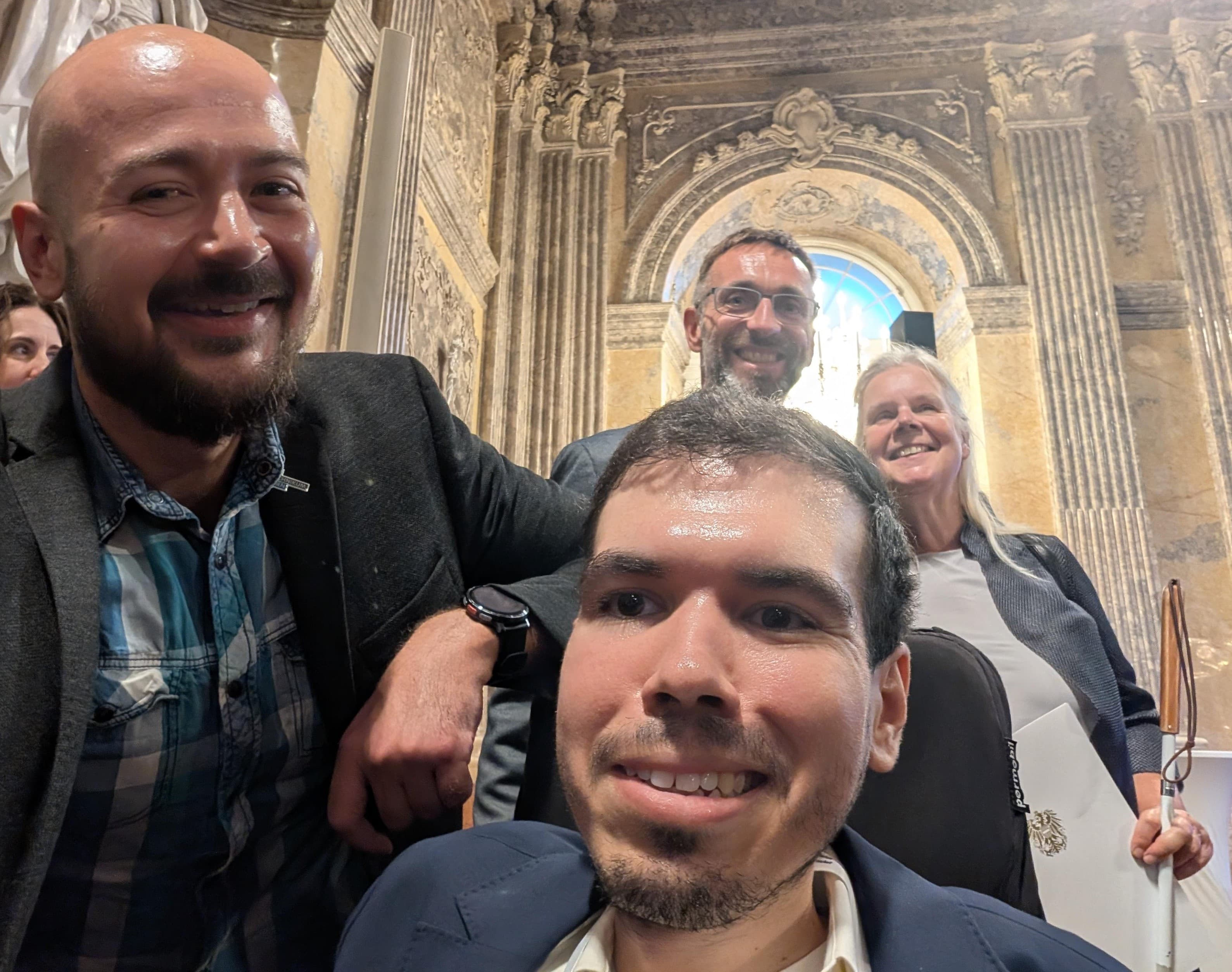 Vier Menschen, die lächeln und für ein Selfie posieren, vor dem Hintergrund eines prunkvollen, historischen Gebäudes mit kunstvoll verzierten Säulen und Bögen.