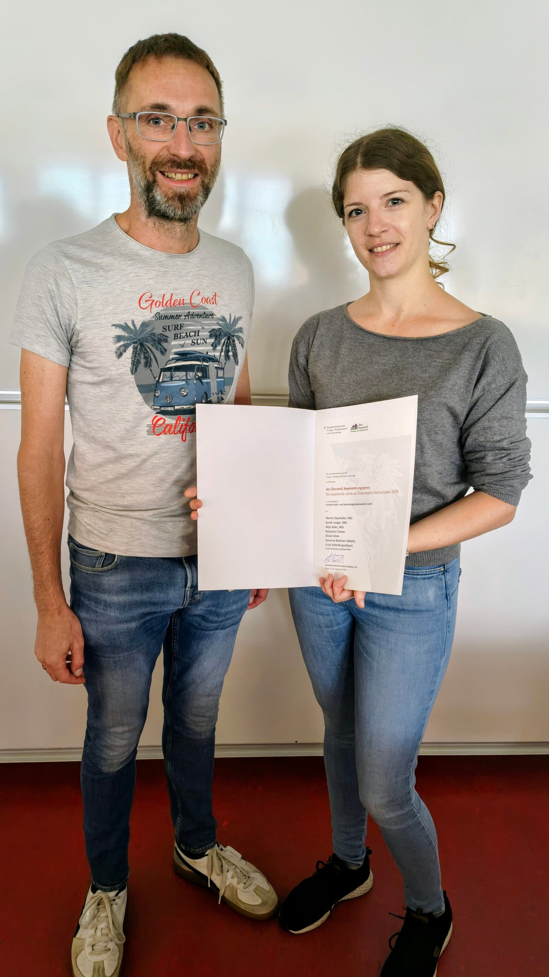Zwei Personen stehen in einem Raum und lächeln in die Kamera. Die Person auf der rechten Seite hält eine geöffnete Mappe in der Hand, in der sich ein Zertifikat befindet. Beide sind leger gekleidet, und im Hintergrund ist ein Whiteboard zu sehen.