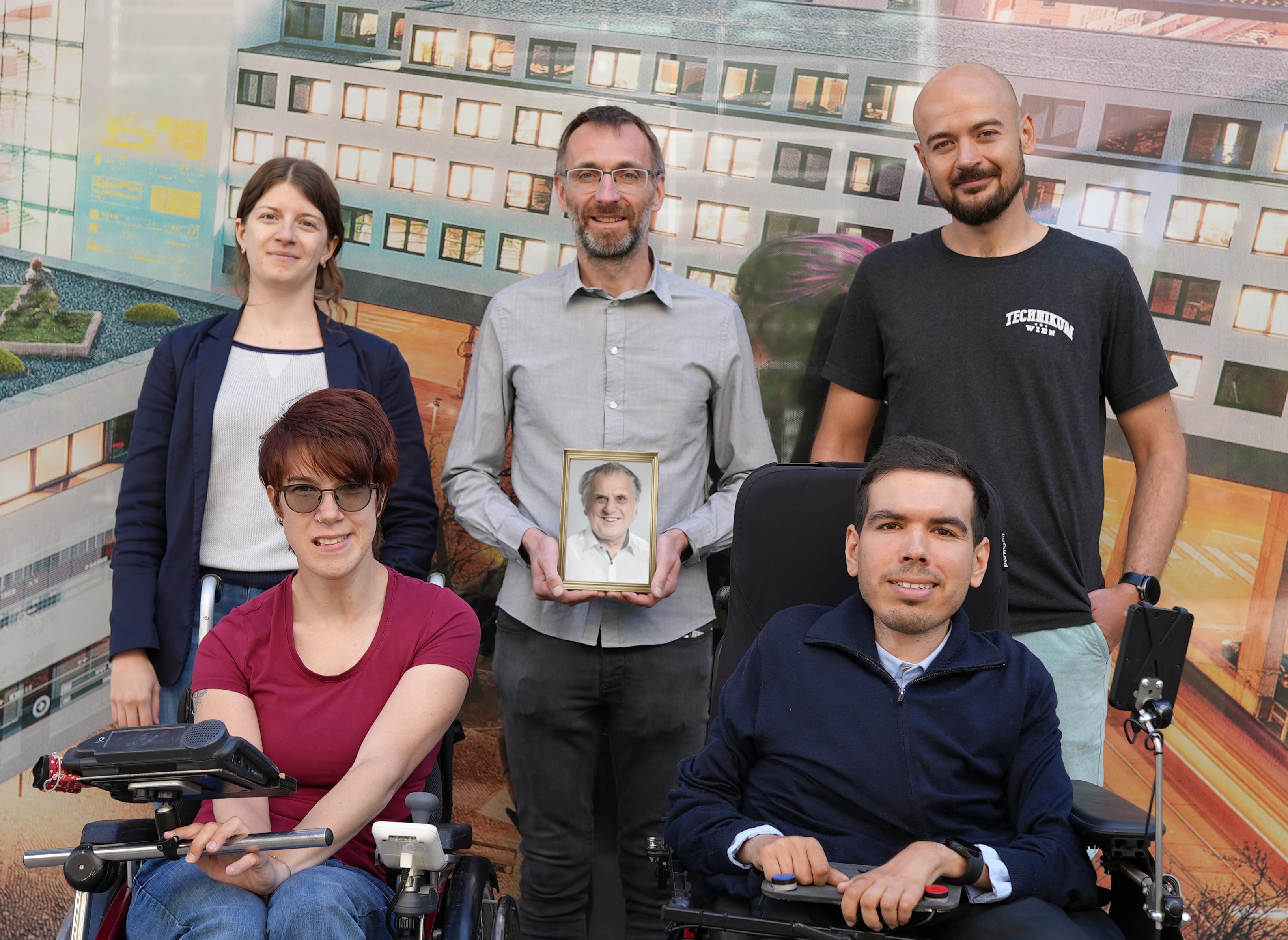Fünf Erwachsene posieren vor einem Wandgemälde. Zwei von ihnen sitzen in Elektrorollstühlen, einer hält ein gerahmtes Foto eines älteren Mannes in der Hand, und alle lächeln in die Kamera.