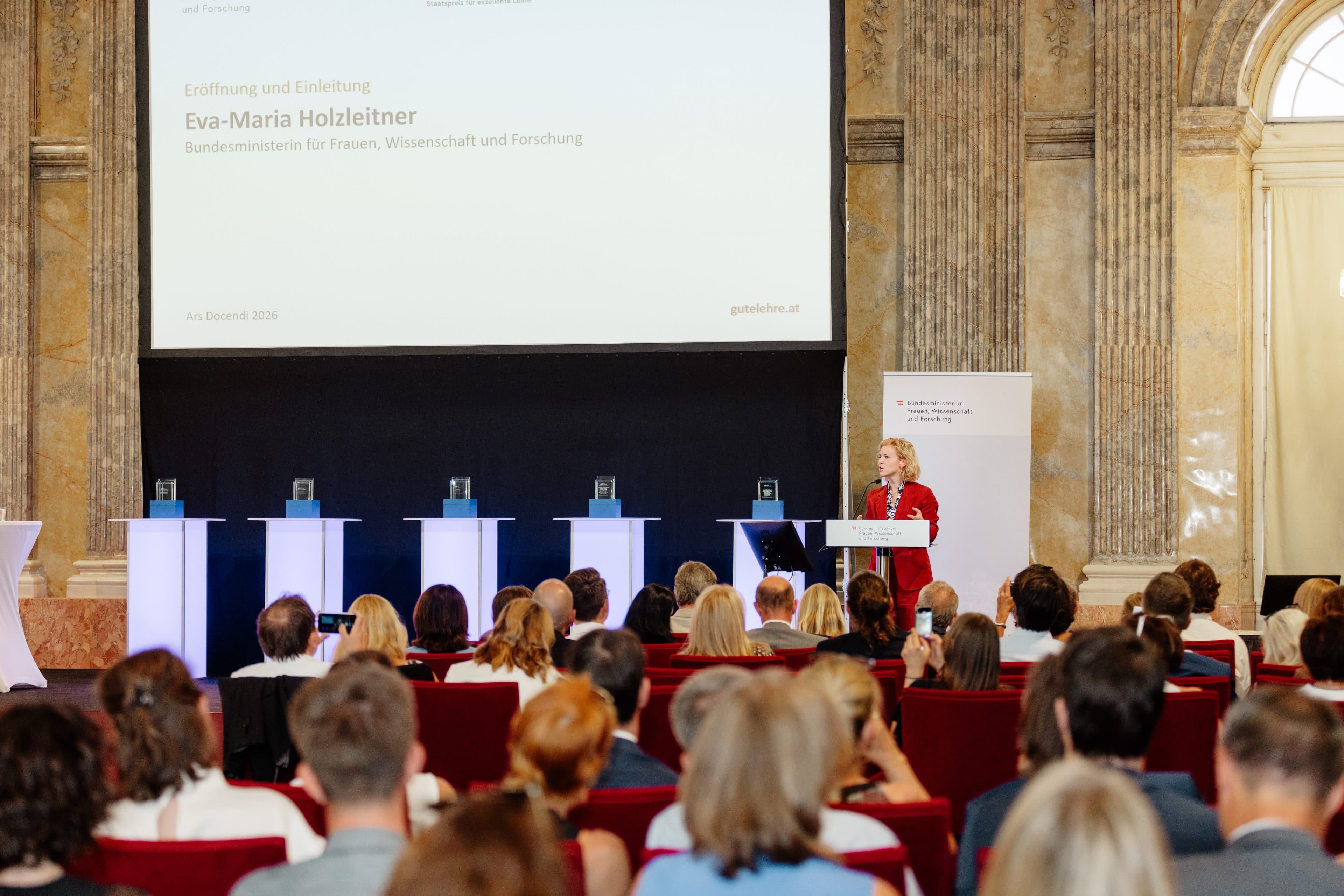 Eine Frau spricht von einem Rednerpult aus vor einem sitzenden Publikum in einem prächtigen Saal mit Marmorsäulen, hinter ihr befinden sich eine große Präsentationsleinwand und ein Banner.