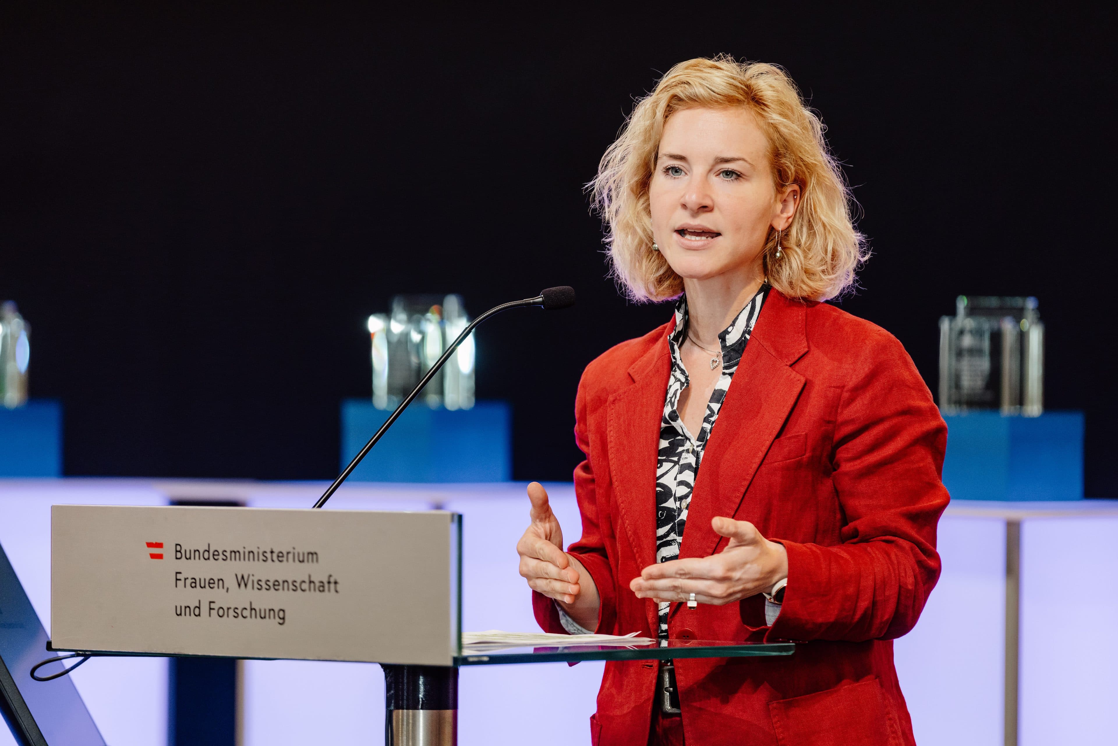 Eine Frau in einem roten Blazer hält eine Rede an einem Rednerpult mit der Aufschrift „Bundesministerium für Frauen, Wissenschaft und Forschung“. Sie gestikuliert mit beiden Händen. Der Hintergrund ist dunkel, im Vordergrund stehen blau beleuchtete Ständer mit Glasobjekten.