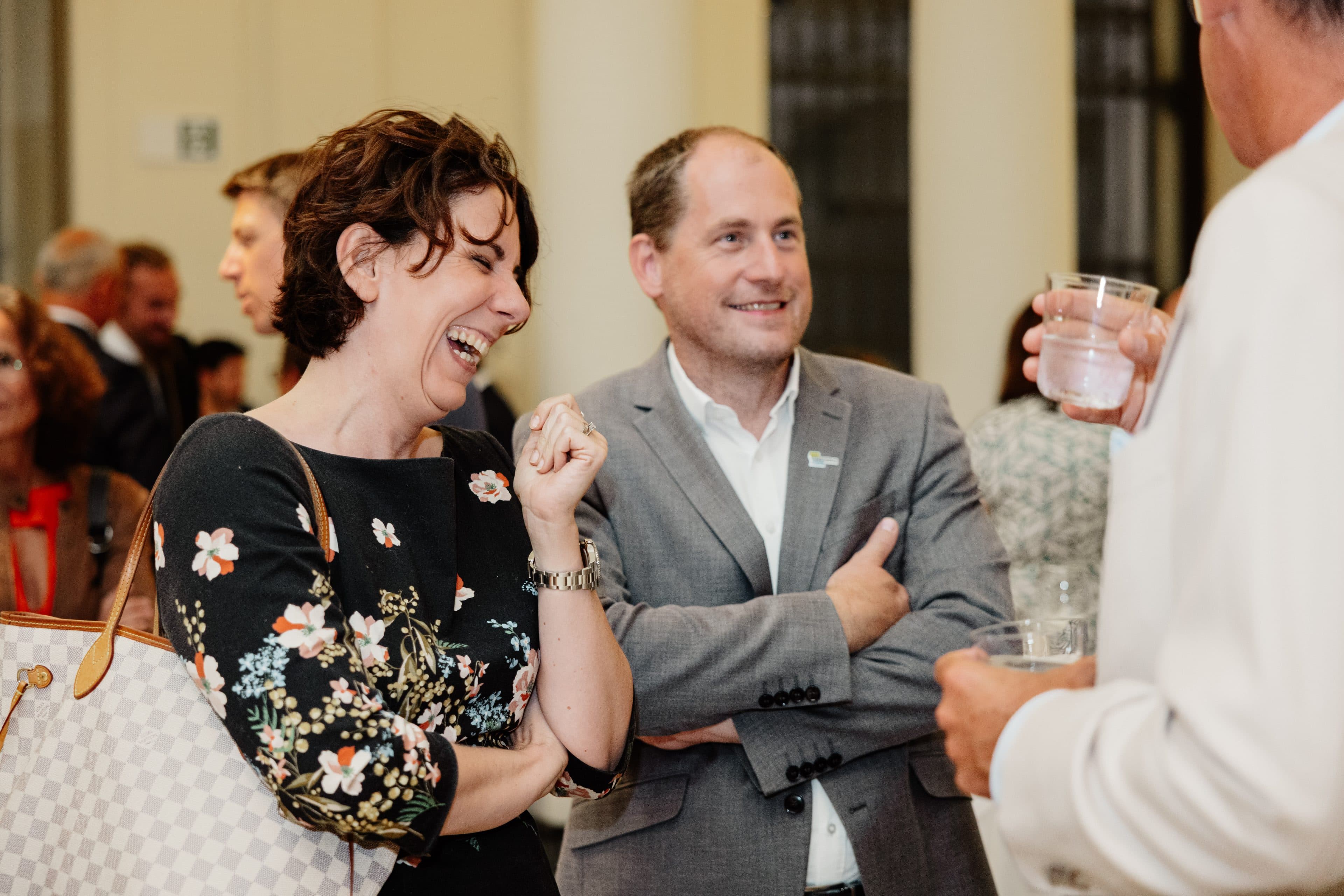 Eine Frau lacht und gestikuliert, während ein Mann neben ihr bei einer gesellschaftlichen Veranstaltung in geschlossenen Räumen lächelt; beide sind in Business-Kleidung gekleidet. Im Hintergrund unterhalten sich weitere Personen miteinander.