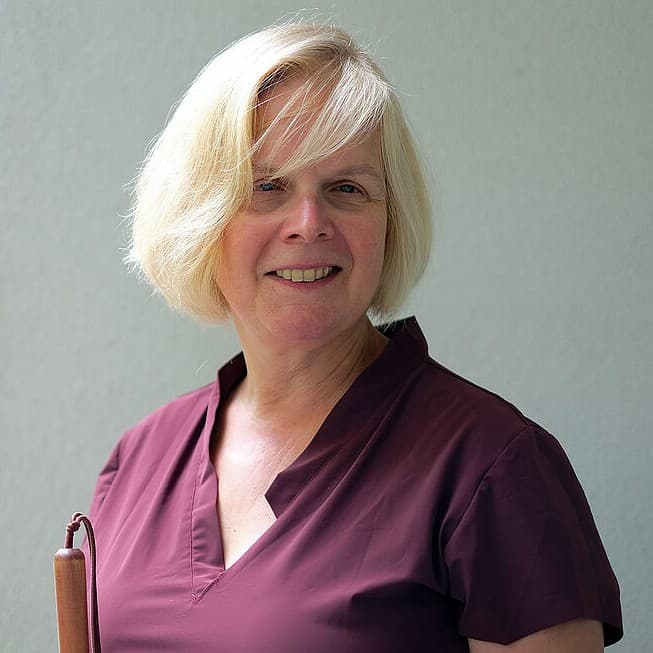 Eine Frau mit kurzen blonden Haaren und einer burgunderroten Bluse steht lächelnd mit einem braunen Gehstock in der Hand vor einem schlichten, hellen Hintergrund.