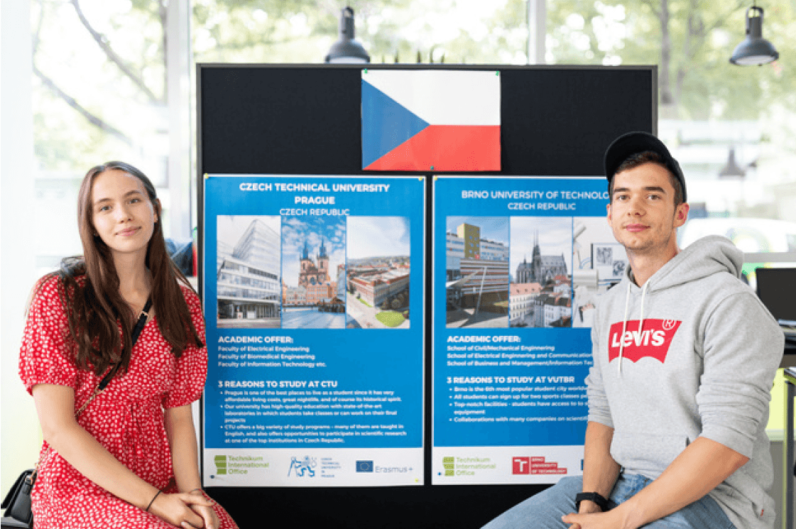 Zwei Studenten sitzen vor Plakaten über tschechische Universitäten, über ihnen weht die tschechische Flagge. Die Frau trägt ein rotes Kleid, der Mann einen grauen Kapuzenpulli und eine Mütze. Beide blicken in die Kamera und lächeln leicht.