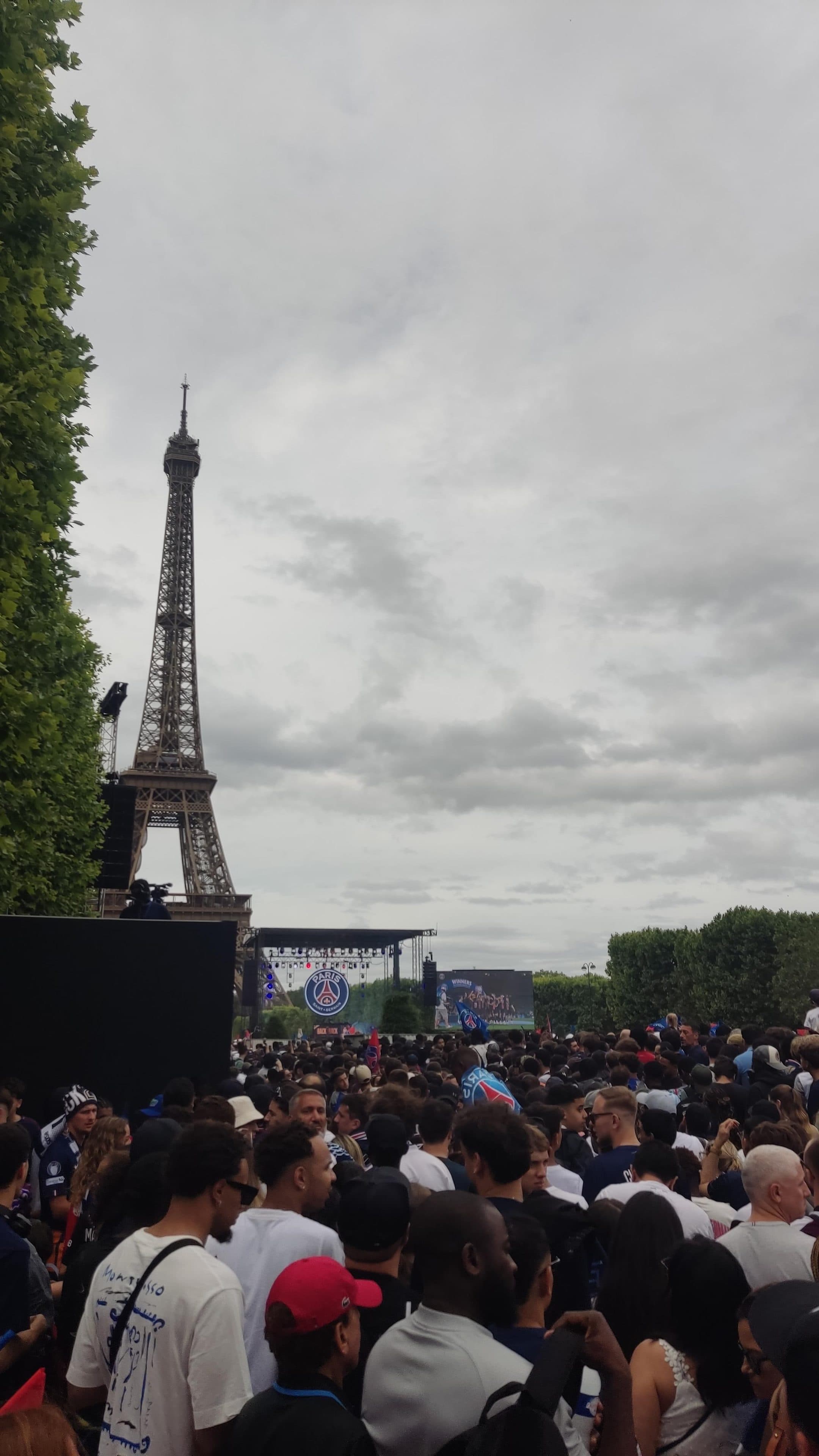In der Nähe des Eiffelturms versammelt sich eine große Menschenmenge vor einer Bühne mit Bildschirmen und Paris-Saint-Germain-Logos (PSG). Der Himmel ist bewölkt, und Bäume rahmen die Szene ein.