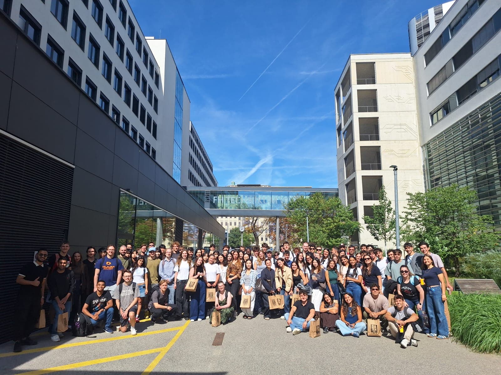 An einem sonnigen Tag posiert eine große Gruppe von Menschen vor modernen Bürogebäuden für ein Foto, im Hintergrund sind Grünflächen und ein blauer Himmel zu sehen.