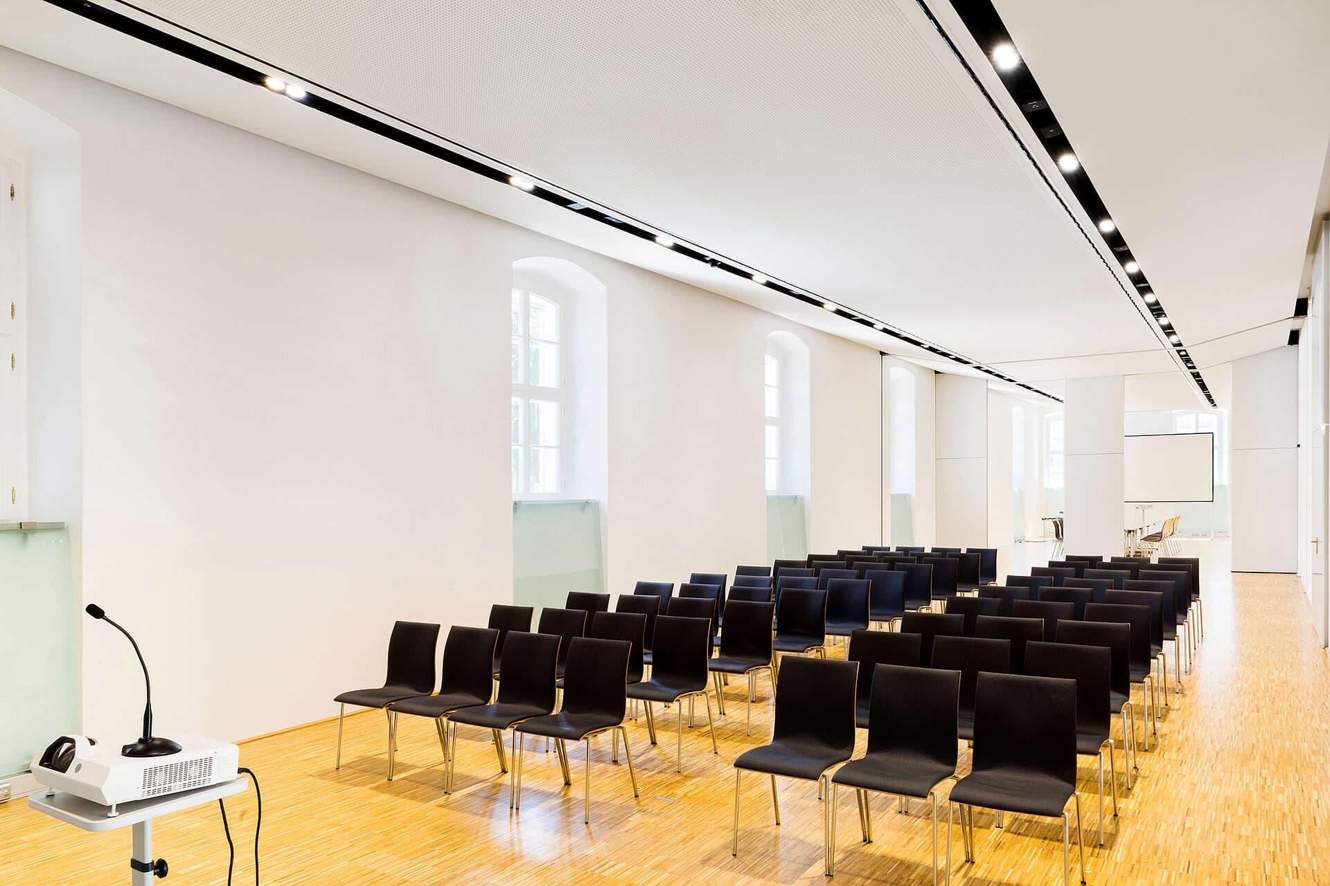 Ein moderner, hell beleuchteter Konferenzraum mit Reihen schwarzer Stühle, die auf einen Projektor und eine Leinwand ausgerichtet sind. An einer Wand reihen sich große Fenster, durch die Tageslicht hereinfällt. Der Boden ist aus Holz, und im Raum befinden sich keine Personen.