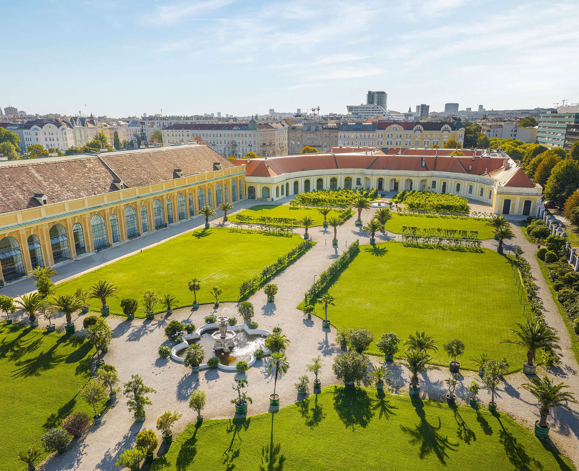 Luftaufnahme eines prächtigen Barockgebäudes mit geschwungener Fassade, großen Fenstern und gepflegten Gärten mit grünen Rasenflächen, Spazierwegen und einem zentralen Springbrunnen unter strahlend blauem Himmel.