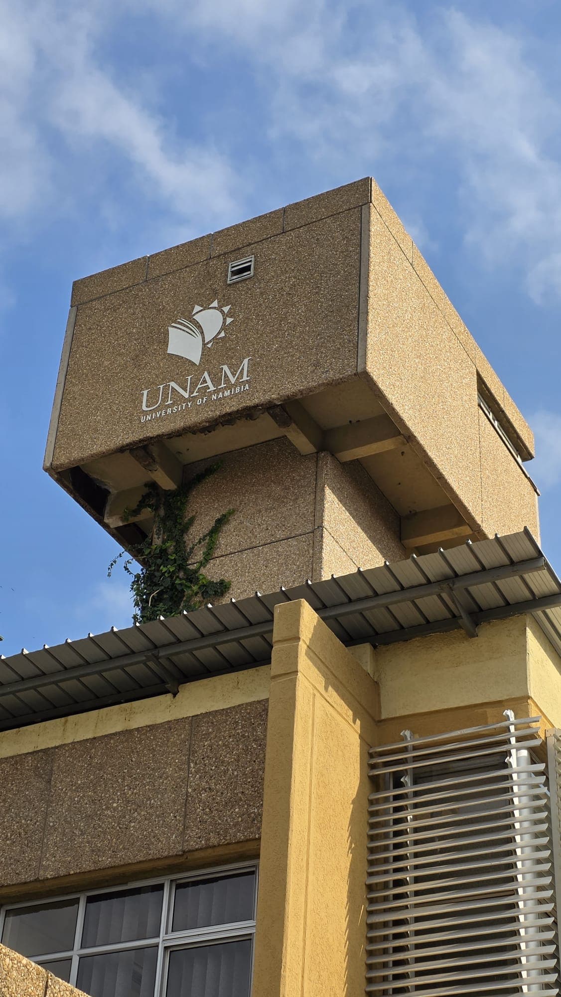 Vor blauem Himmel ist ein braunes Betongebäude mit dem UNAM-Logo (Universität von Namibia) und einer Kletterpflanze zu sehen.