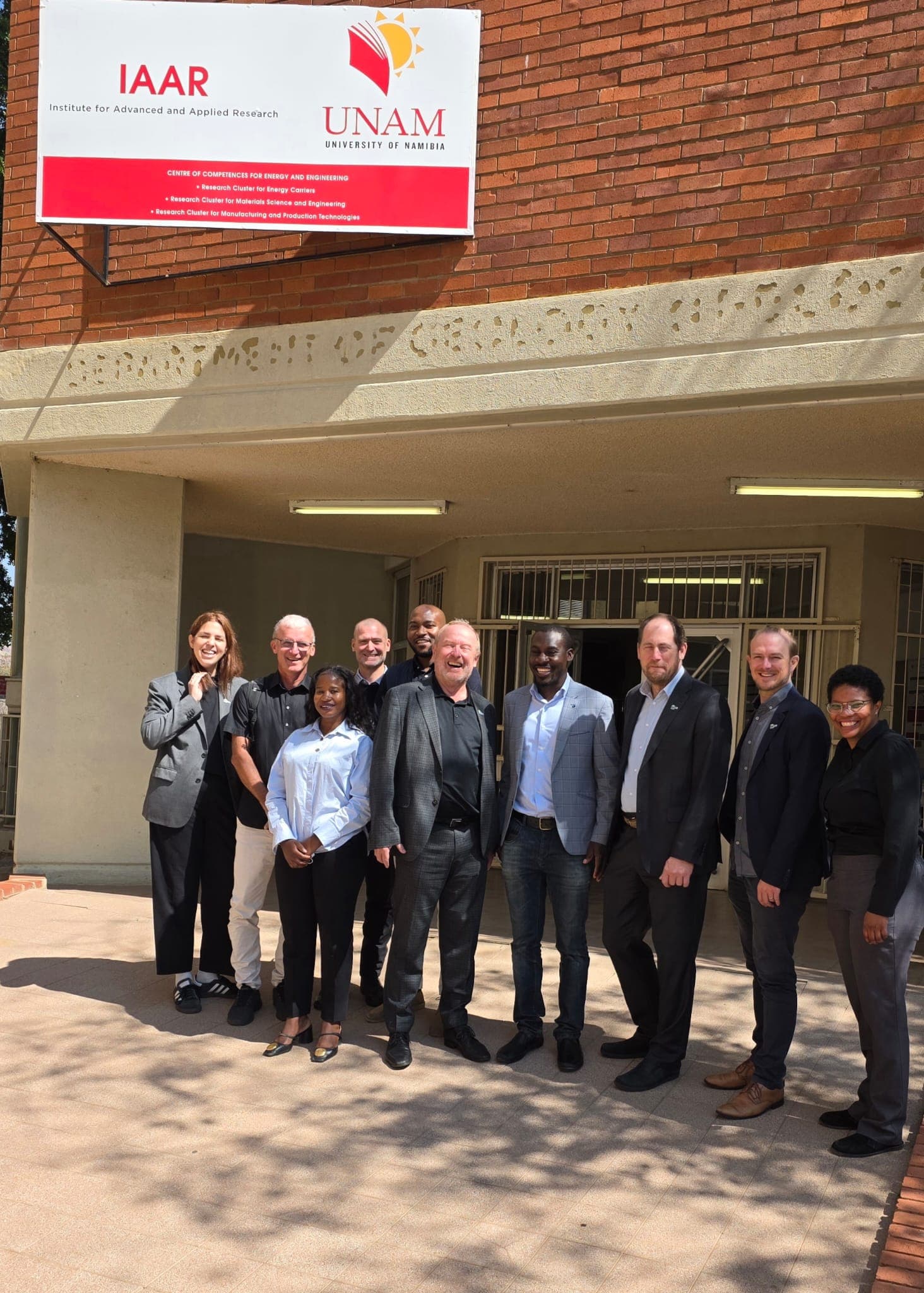 Eine Gruppe von zehn Personen in Geschäftskleidung steht lächelnd vor einem Gebäude unter einem Schild mit der Aufschrift „IAAR“ und „UNAM University of Namibia“.