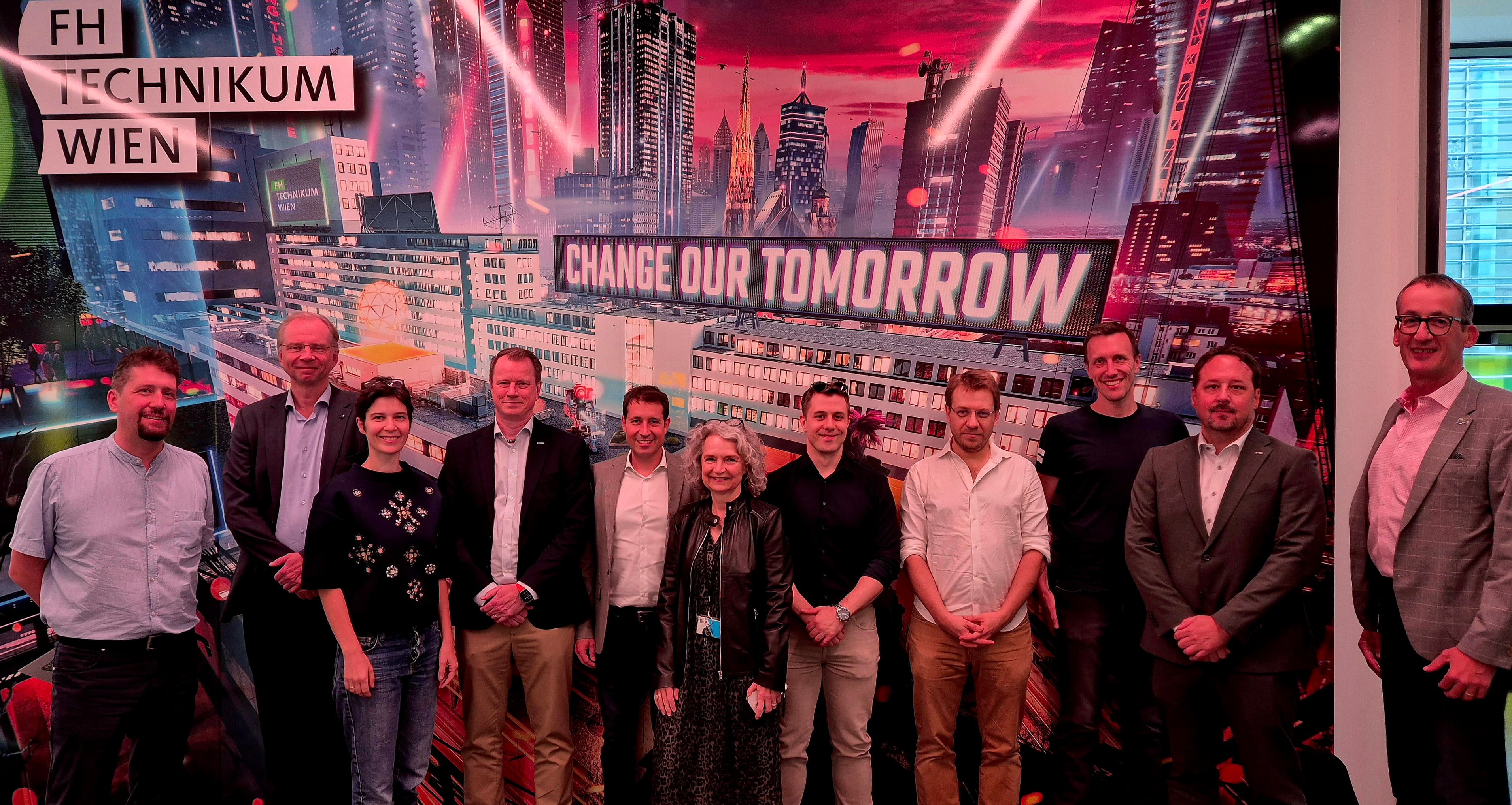 Elf Personen stehen vor einer farbenfrohen Stadtkulisse, auf der die Schriftzüge „CHANGE OUR TOMORROW“ und „FH TECHNIKUM WIEN“ zu sehen sind. Sie lächeln und blicken in die Kamera.