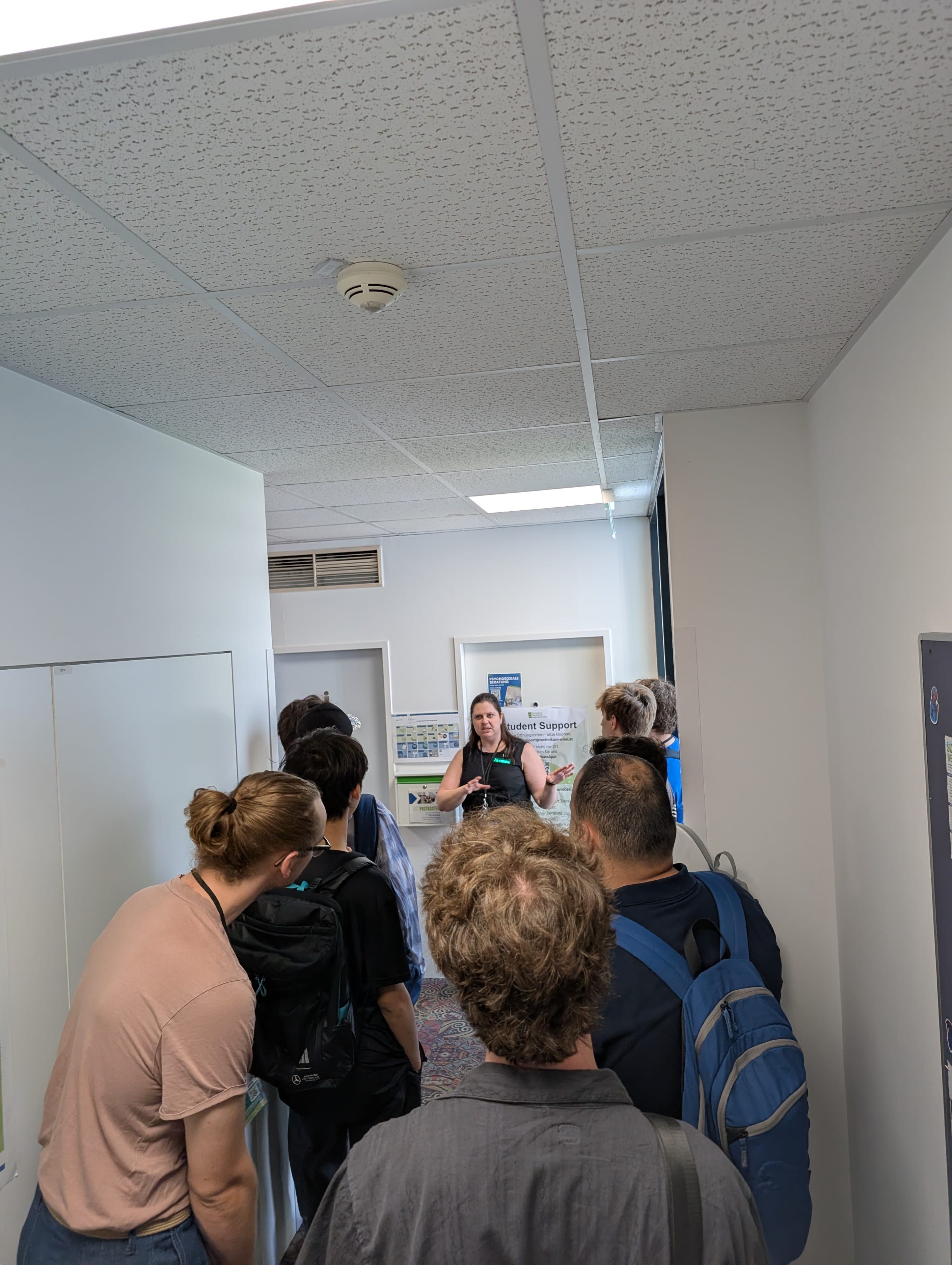 Eine Gruppe von Menschen steht in einem Flur und hört einer Frau zu, die vor ihnen spricht. Der Flur hat weiße Wände, eine gesprenkelte Decke und Bürotüren. Einige der Anwesenden tragen Rucksäcke.