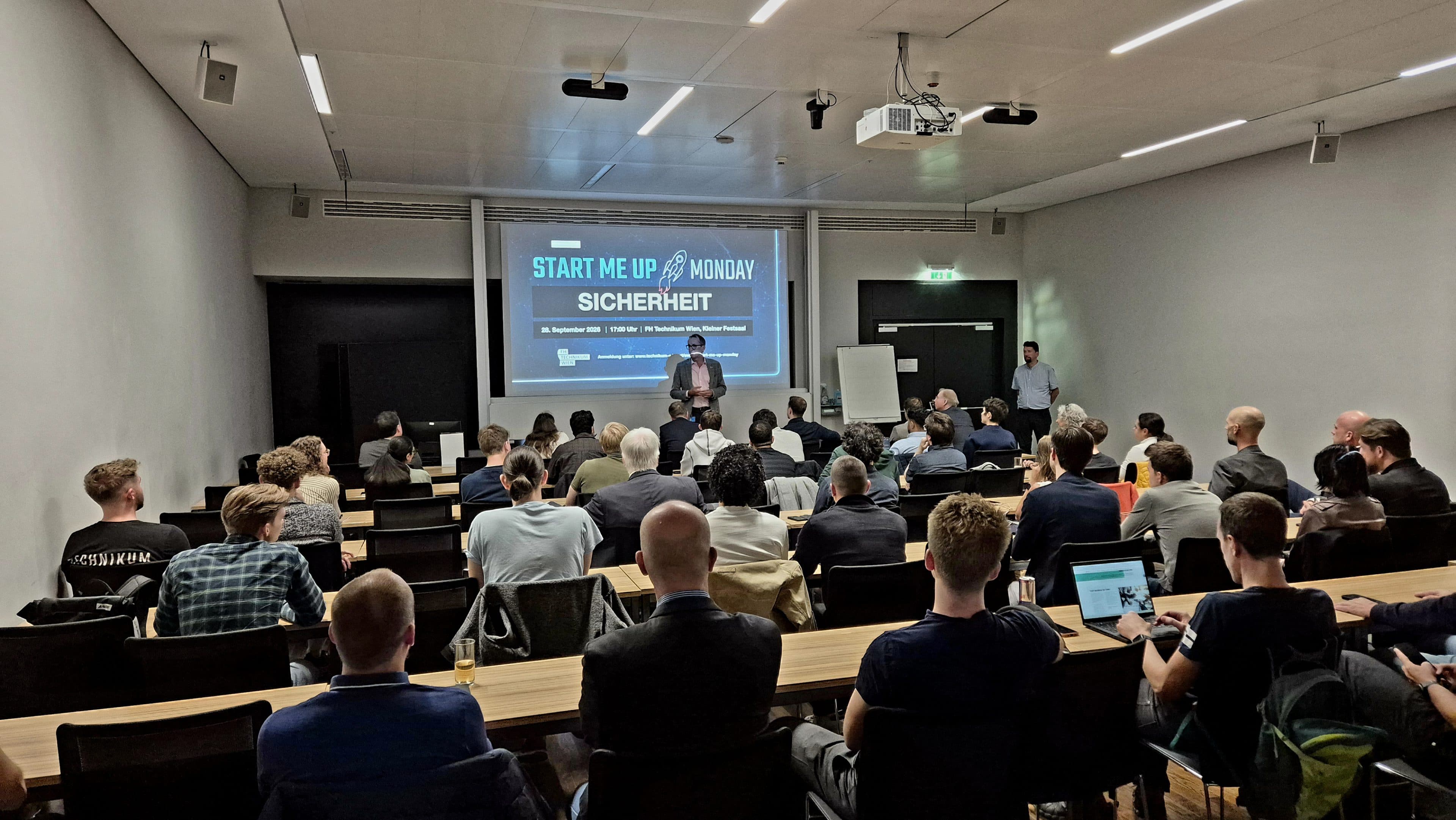 Eine Gruppe von Menschen sitzt in einem modernen Konferenzraum und hört einem Redner zu, der vorne steht. Auf einer großen Leinwand wird eine Präsentation zum Thema „Sicherheit“ gezeigt. Die meisten Teilnehmer blicken nach vorne, einige arbeiten an ihren Laptops.
