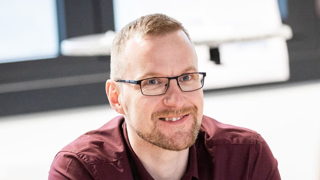 Ein Mann mit kurzen hellbraunen Haaren, Bart und Brille lächelt und trägt ein weinrotes Hemd. Der Hintergrund ist hell und unscharf.