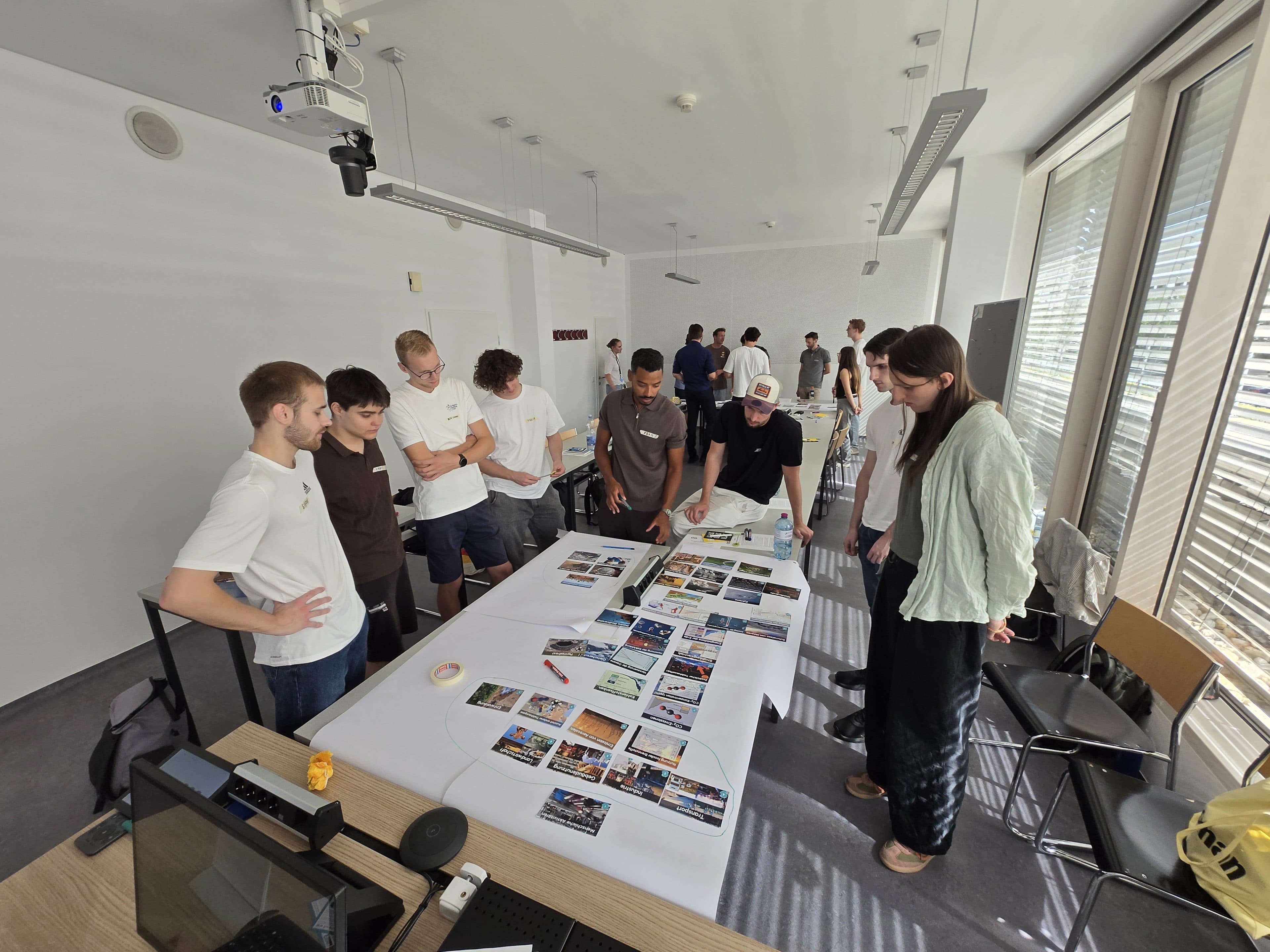 Eine Gruppe von Menschen steht um einen Tisch herum, der mit großen Blättern und verschiedenen Fotos bedeckt ist, und diskutiert und arbeitet gemeinsam in einem hellen, modernen Besprechungsraum.