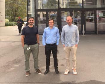 Drei Männer stehen vor einem modernen Glasgebäude. Zwei tragen Hemden und Hosen, während einer ein T-Shirt und eine Hose trägt. Im Hintergrund sind Bäume und eine weitere Person zu sehen.