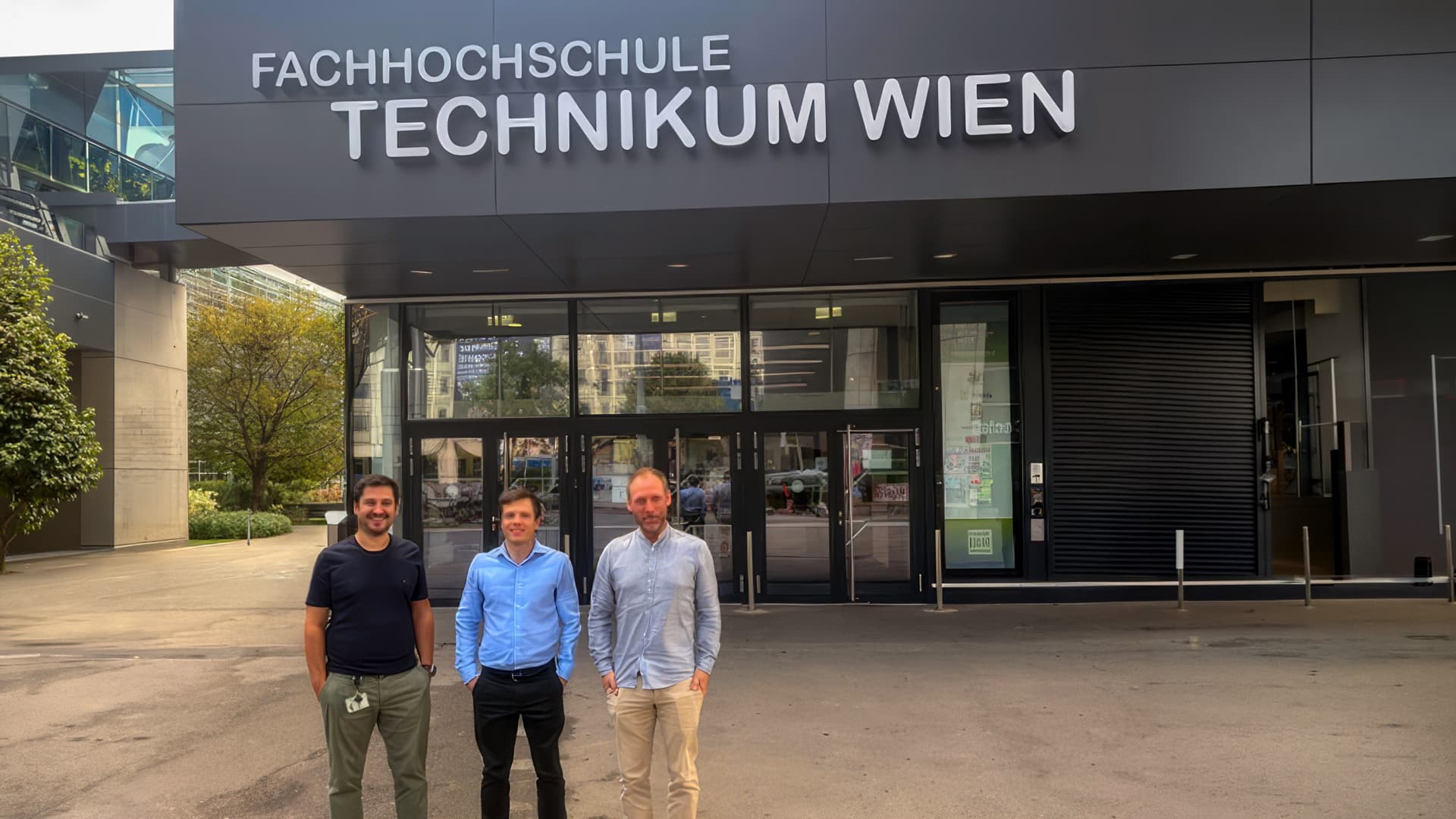 Drei Personen stehen vor dem Gebäude der Fachhochschule Technikum Wien, einer modernen Bildungseinrichtung mit großen Glastüren und einem auffälligen Schild über dem Eingang.
