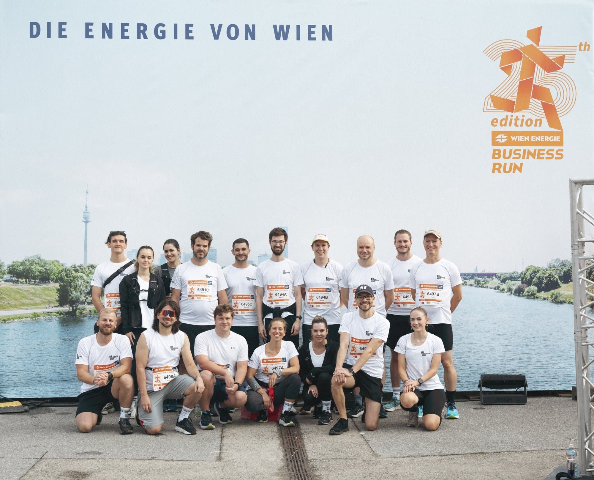 Eine Gruppe von Menschen in einheitlichen weißen Hemden posiert gemeinsam im Freien vor einem Banner für den Wien Energie Business Run in der Nähe eines Flusses.