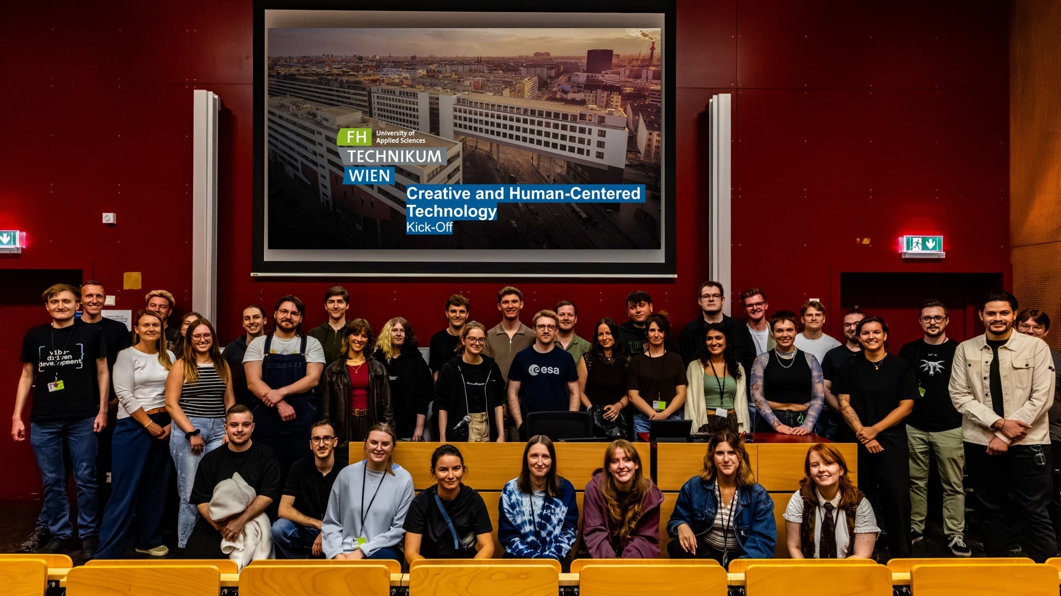 Eine Gruppe lächelnder Menschen posiert in einem Hörsaal vor einer Leinwand, auf der Kick-off zur kreativen und menschenzentrierten Technologie an der FH Technikum Wien zu lesen ist.