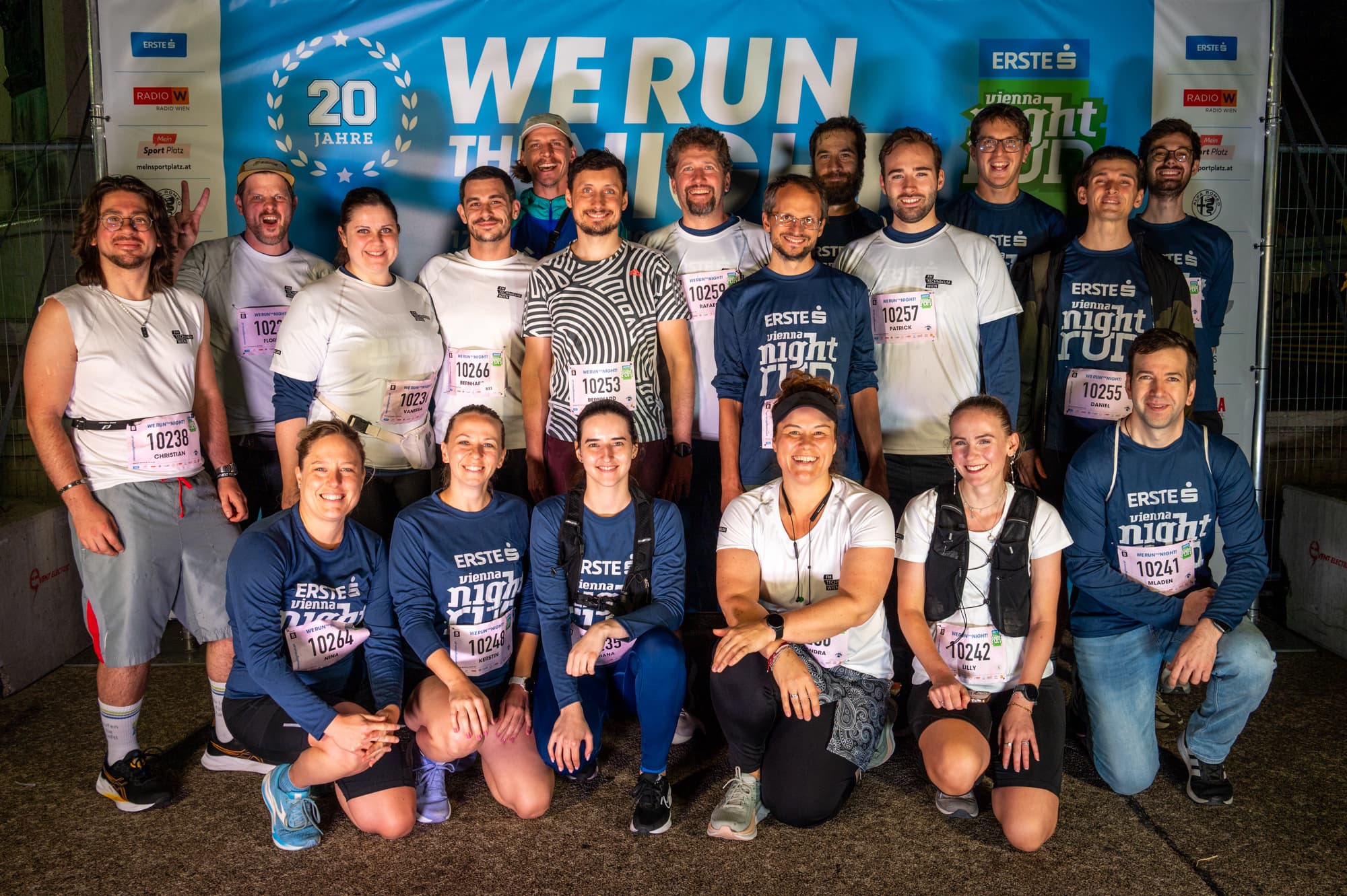 Eine große Gruppe von Läufern mit Startnummern posiert für ein Foto vor einem blauen We Run The Night-Banner, das das 20-jährige Jubiläum feiert. Die Gruppe, bestehend aus Männern und Frauen unterschiedlichen Alters, lächelt, während sie nachts auf einer gepflasterten Fläche stehen und knien.