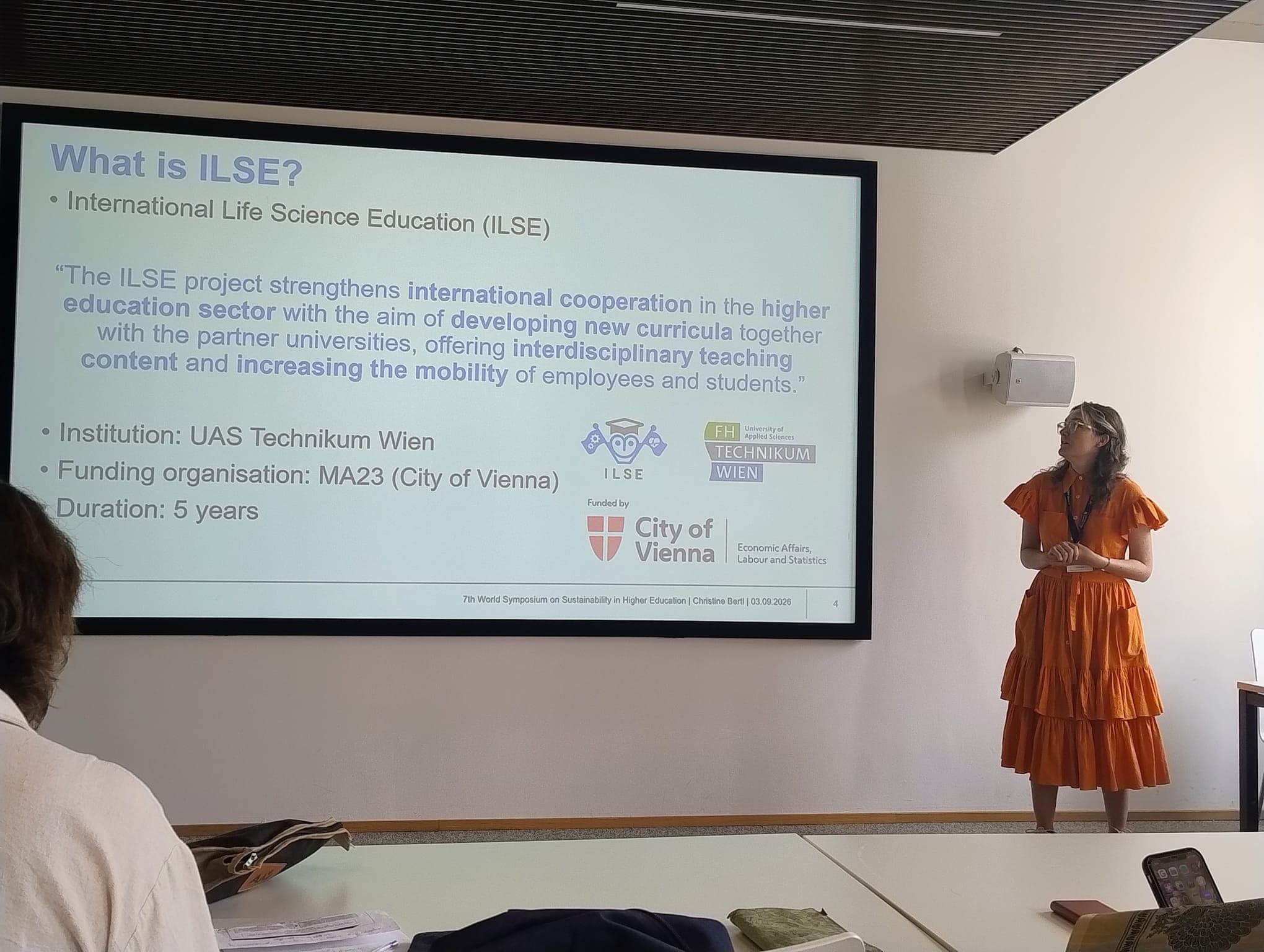 Eine Frau in einem orangefarbenen Kleid hält einen Vortrag in einem Klassenzimmer. Auf der Leinwand werden Informationen zum ILSE-Projekt, zur internationalen Zusammenarbeit und zum Bildungswesen angezeigt, zusammen mit den Logos der Fachhochschule Technikum Wien und der Stadt Wien.